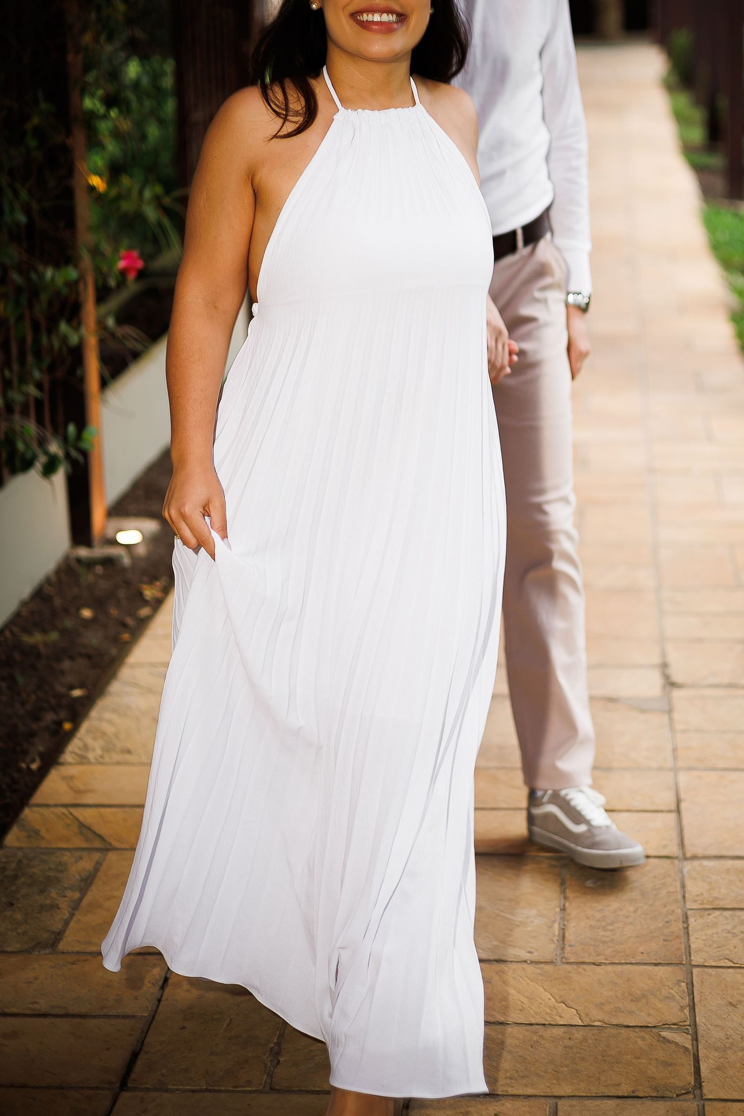Woman smiling in elegant white halter neck pleated maxi dress, standing on stone pathway with greenery.