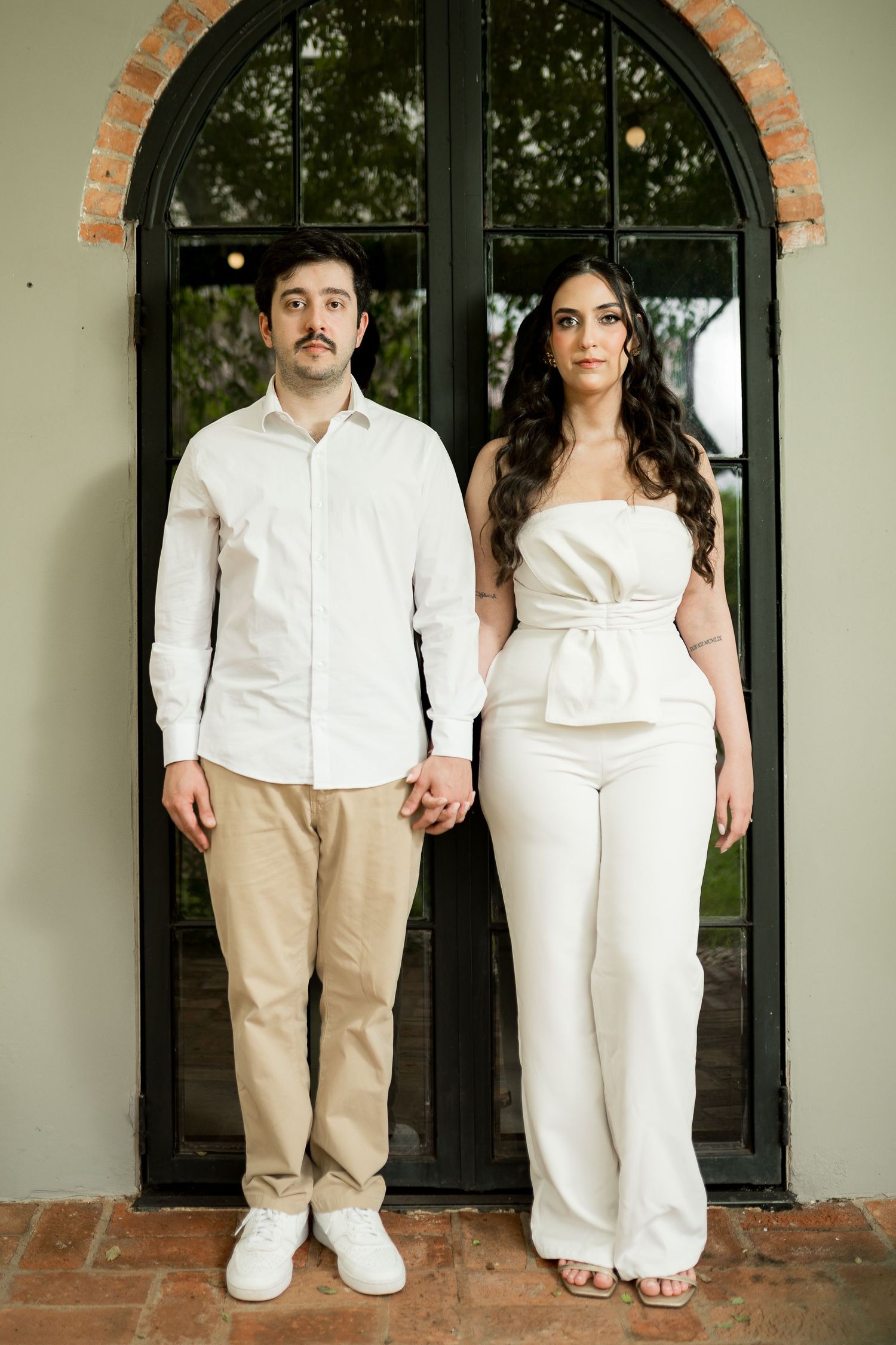 A couple in white outfits holds hands in front of an arched black door, posing for an engagement photo.