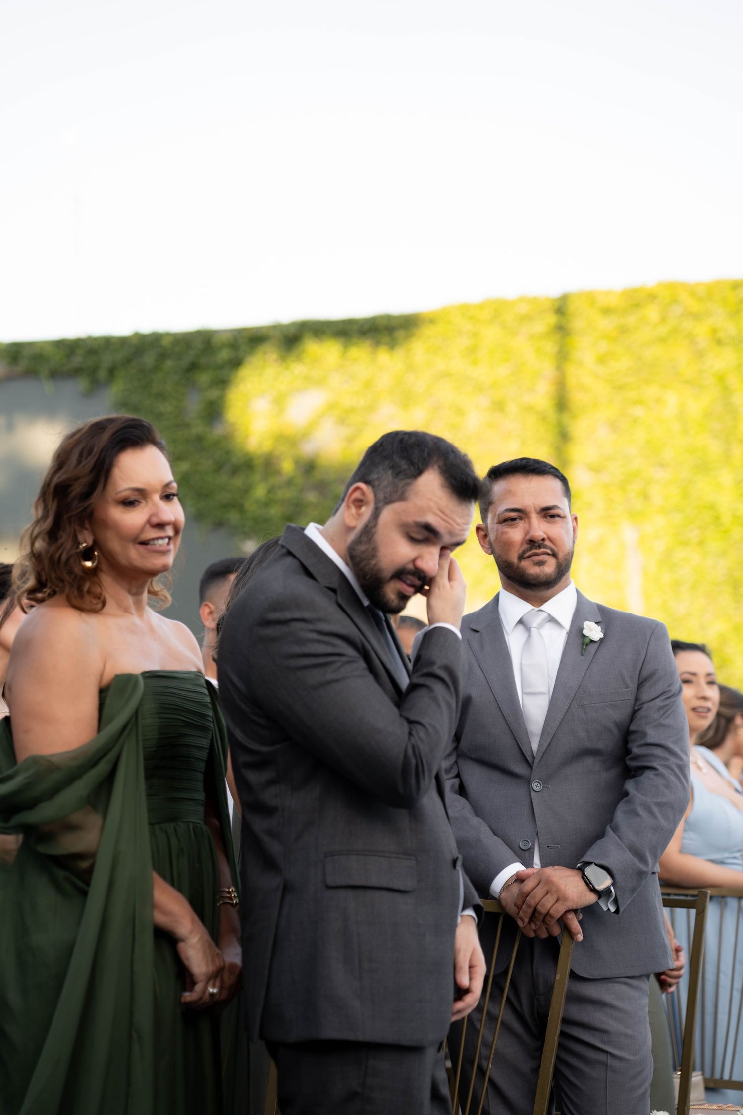 Emotional groom wiping tears at outdoor wedding ceremony, flanked by groomsman and smiling woman in green gown.