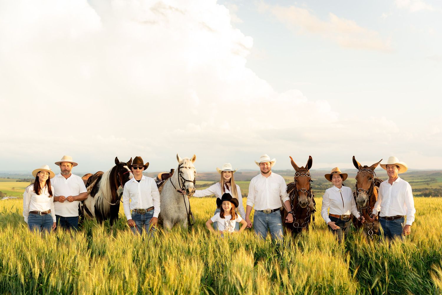 Family in white shirts and cowboy hats standing with horses in a lush green field under a cloudy sky.