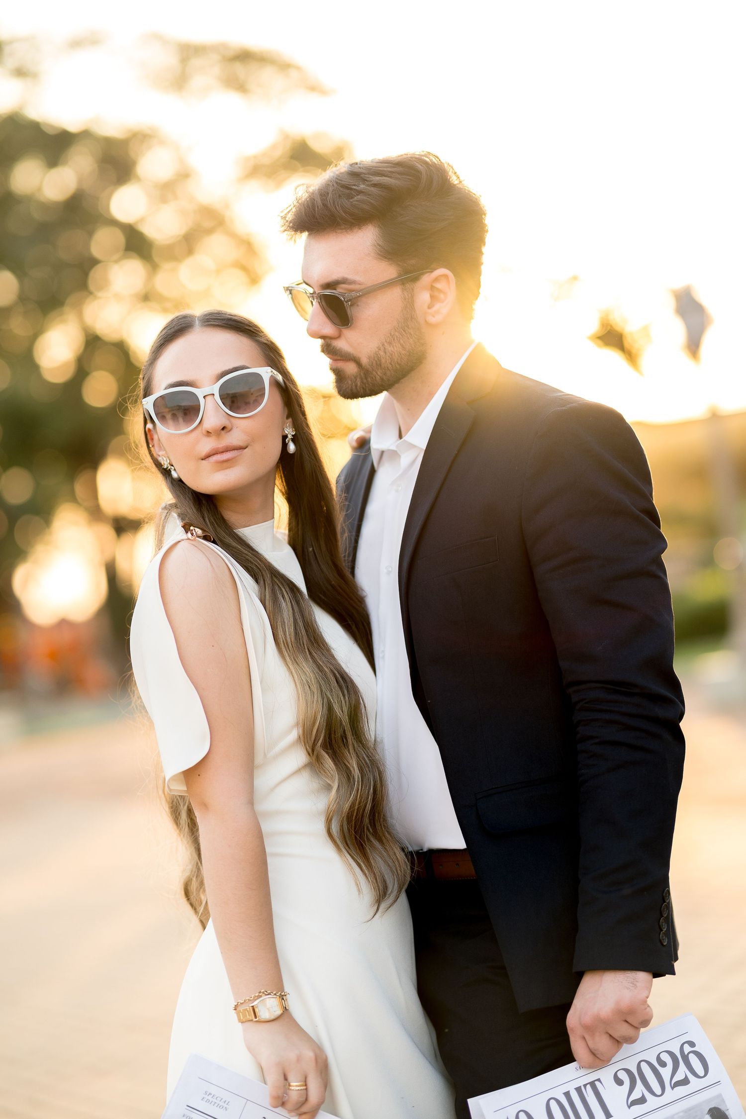 Elegant couple in sunglasses posing at sunset, woman in white dress, man in black suit, holding papers outdoors.