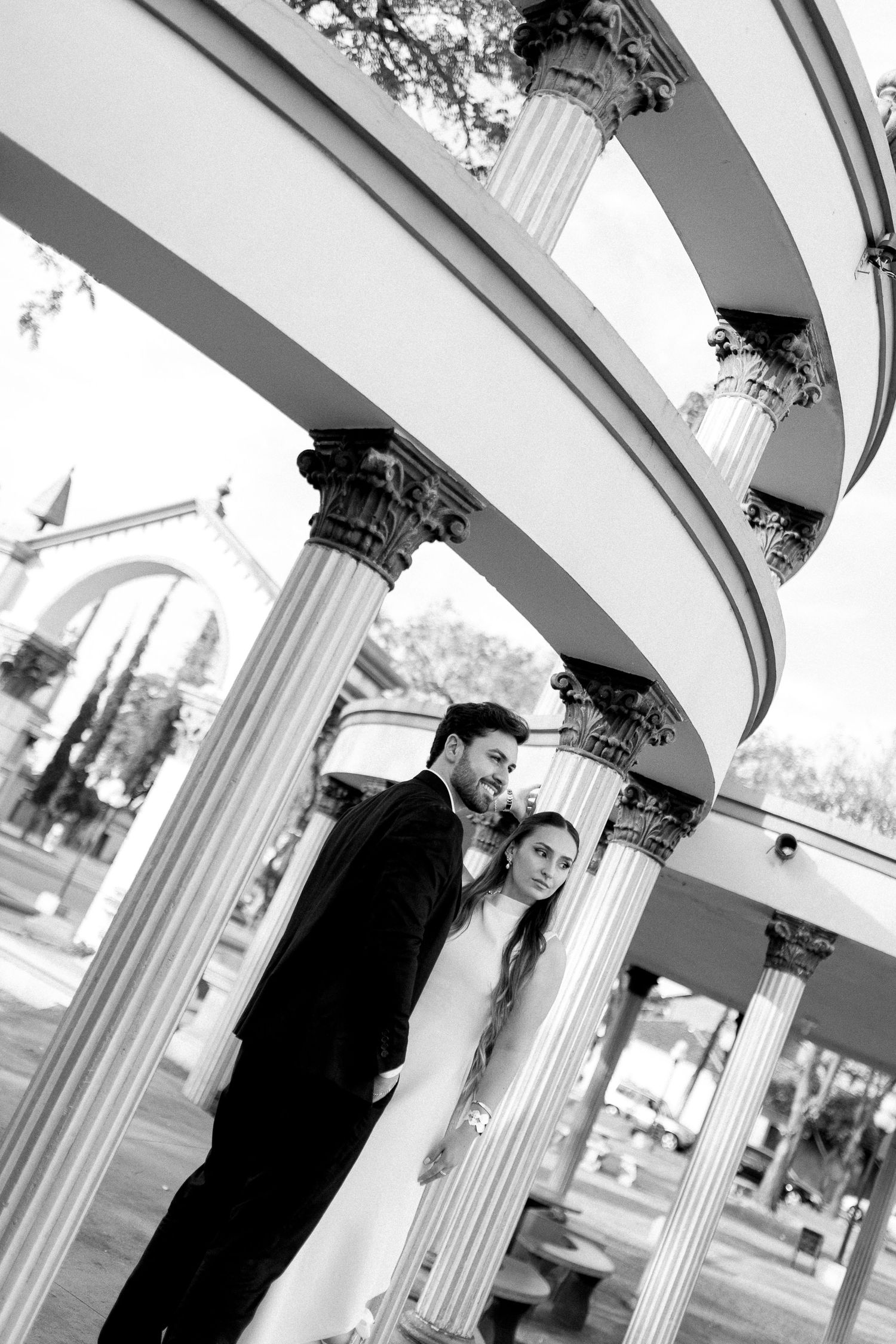 Black and white wedding photo of a couple embracing under elegant spiral colonnade with classical pillars.