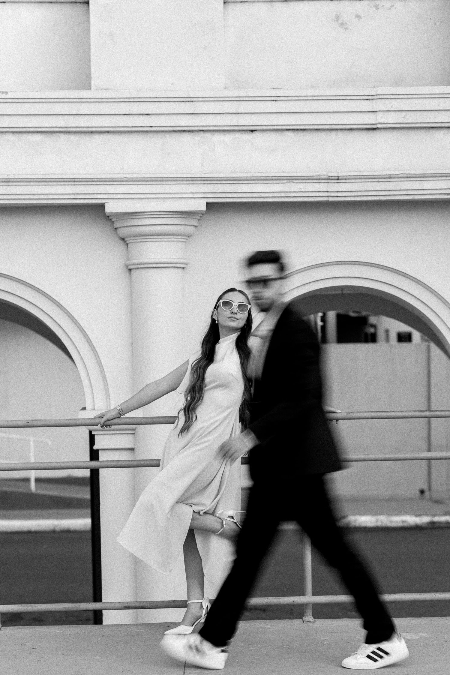 Black and white photo of a woman in a white dress leaning on a railing while a blurred man walks past.