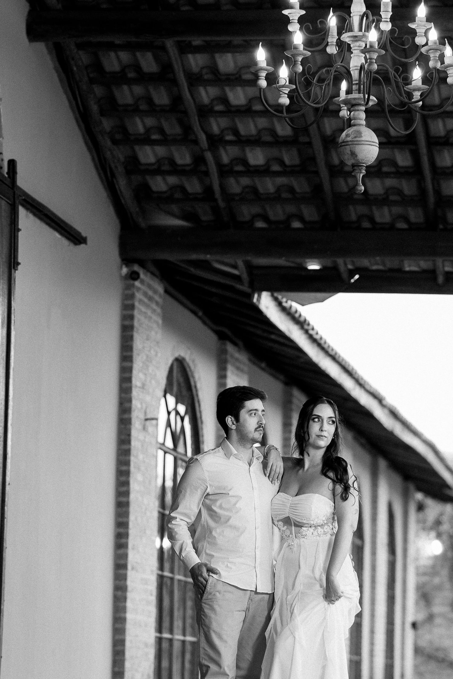Black and white photo of an elegant couple standing on a colonial-style veranda beneath a chandelier.