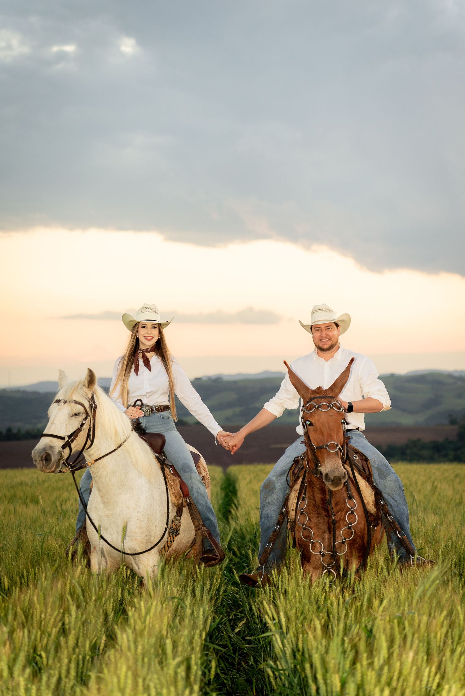 Couple in white shirts and cowboy hats holding hands while riding horses through a green field at sunset.