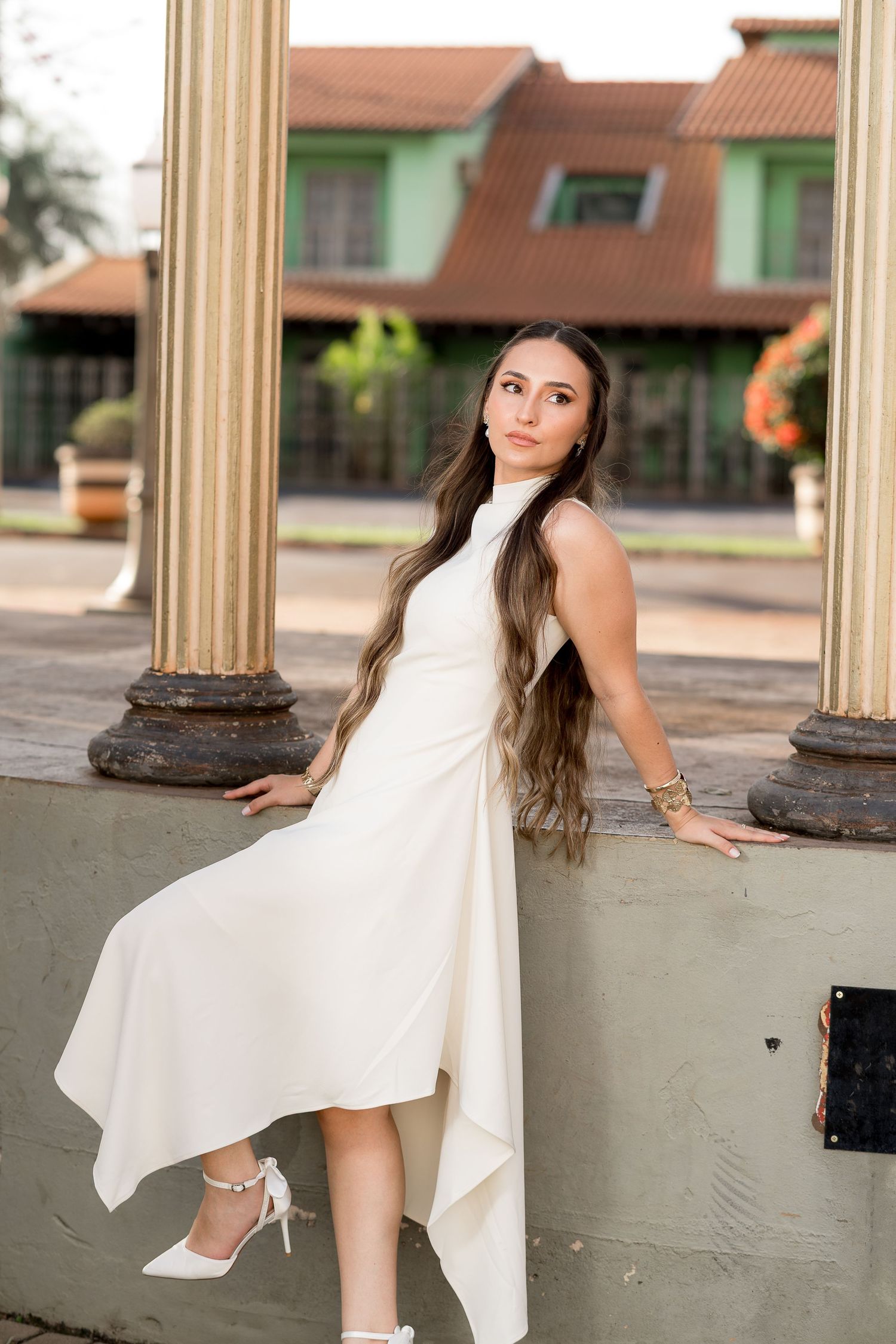 Young woman in elegant white dress and heels poses gracefully against stone columns with green building backdrop.