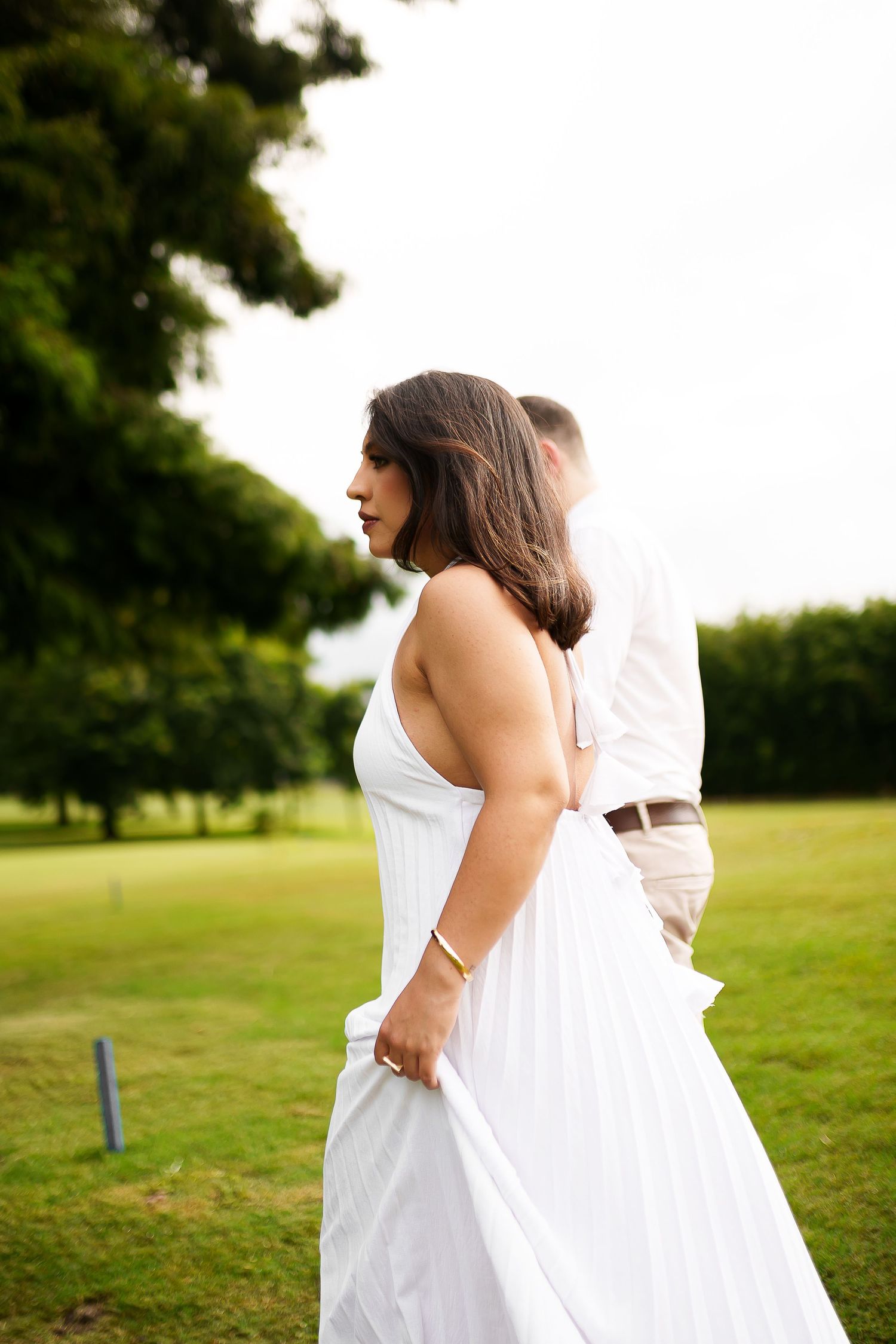 A woman in a white dress stands with a man in a park setting during what appears to be a wedding or engagement shoot.