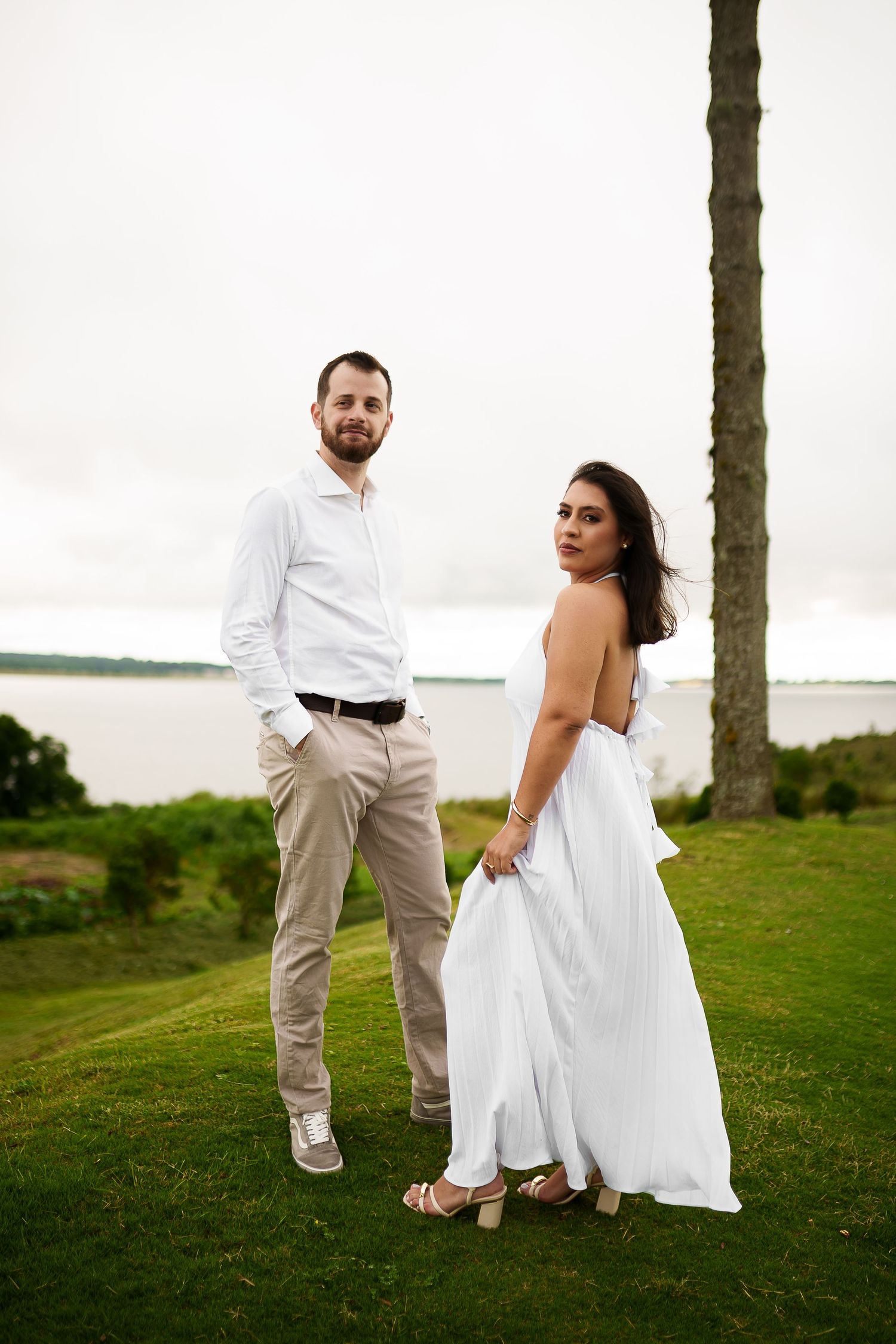 Couple in white outfits posing on green lawn near palm tree with water view on cloudy day.
