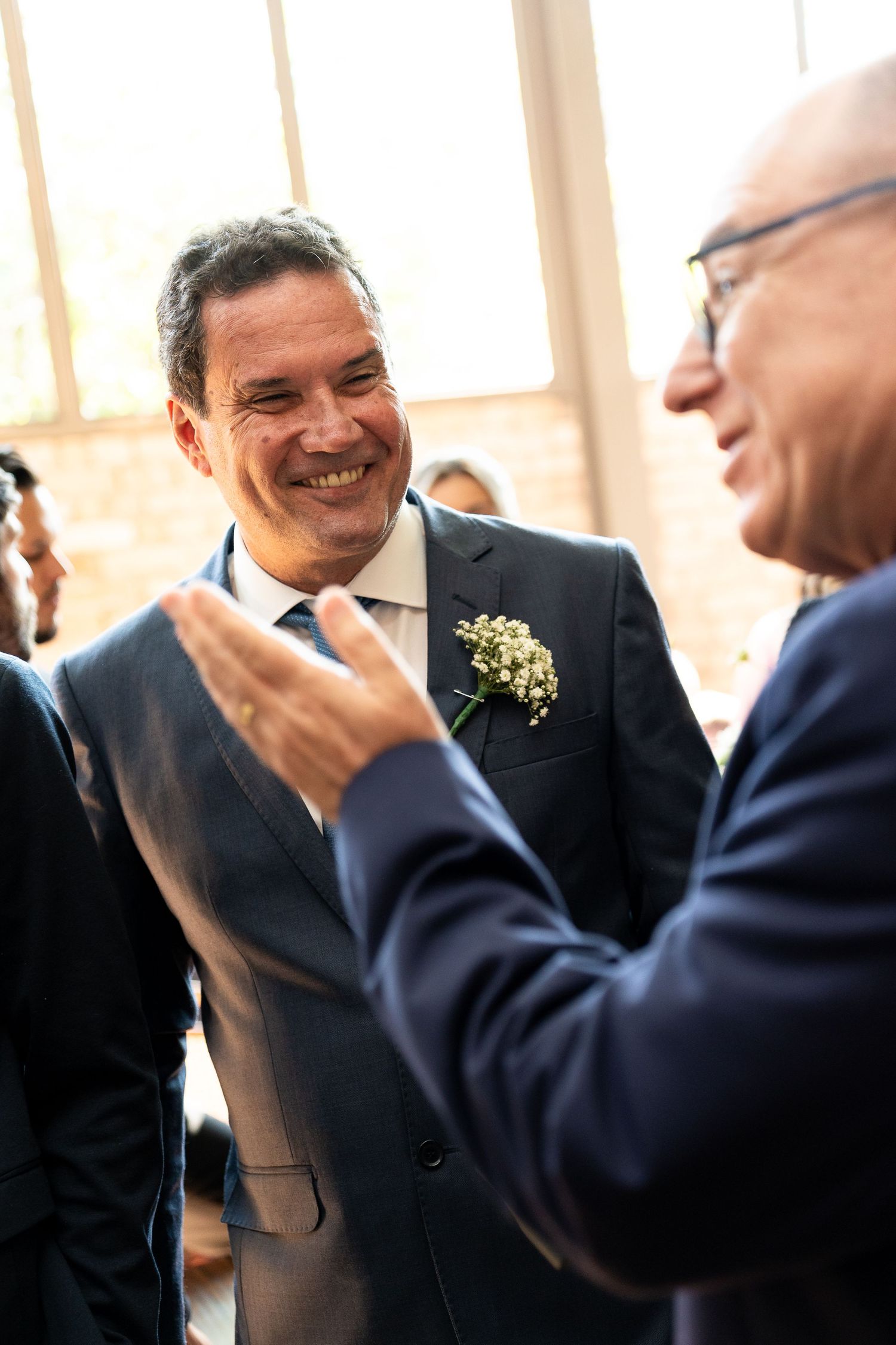 Smiling groom in navy suit with boutonniere receiving congratulations at wedding reception.