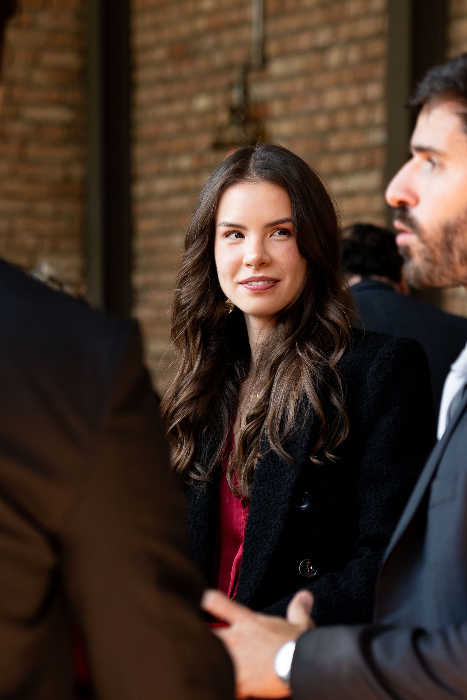 Young brunette woman in black blazer smiling at an outdoor event with brick wall background.