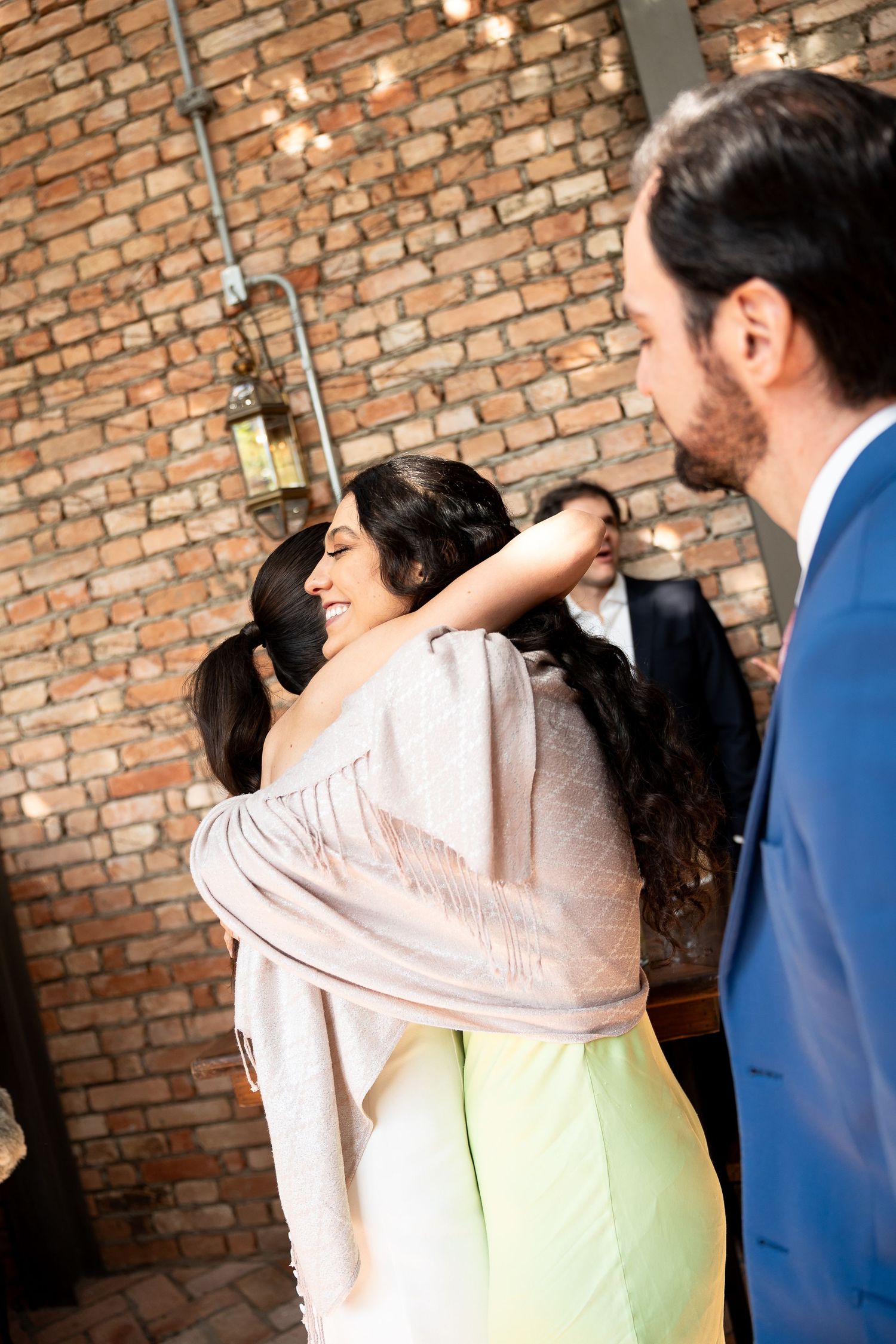 Two women share a warm hug at an indoor event, with a brick wall and string lights in the background.