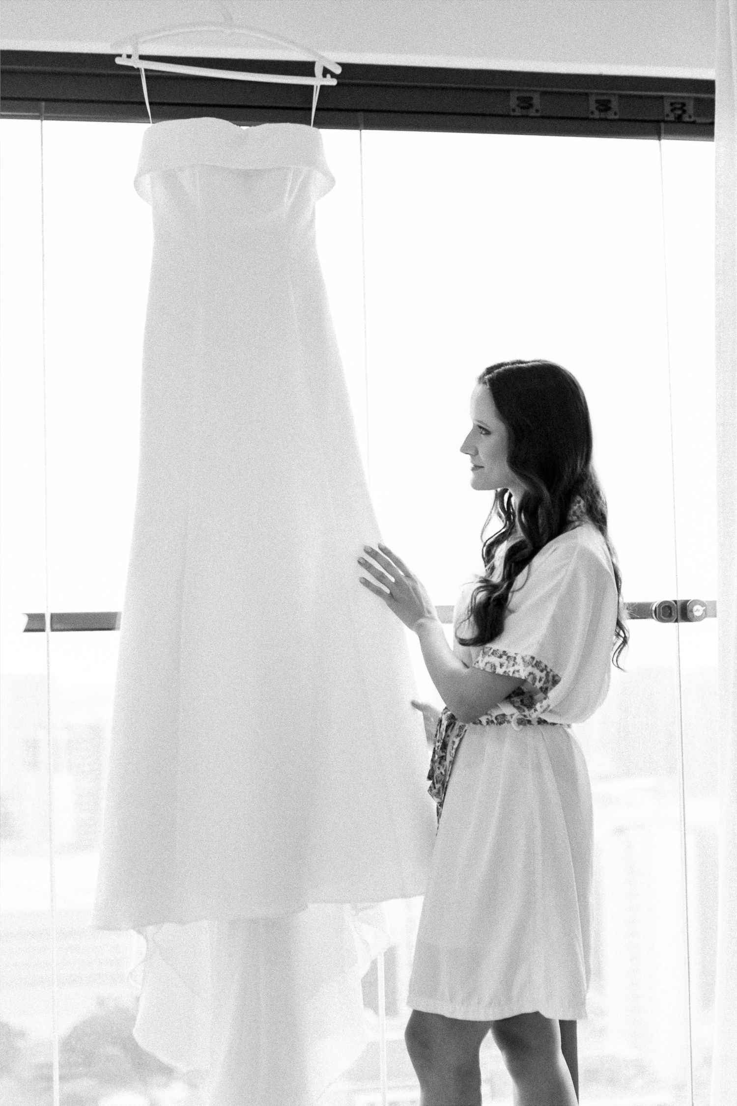 Bride in white robe admiring her wedding gown hanging by a bright window in a black and white photo.