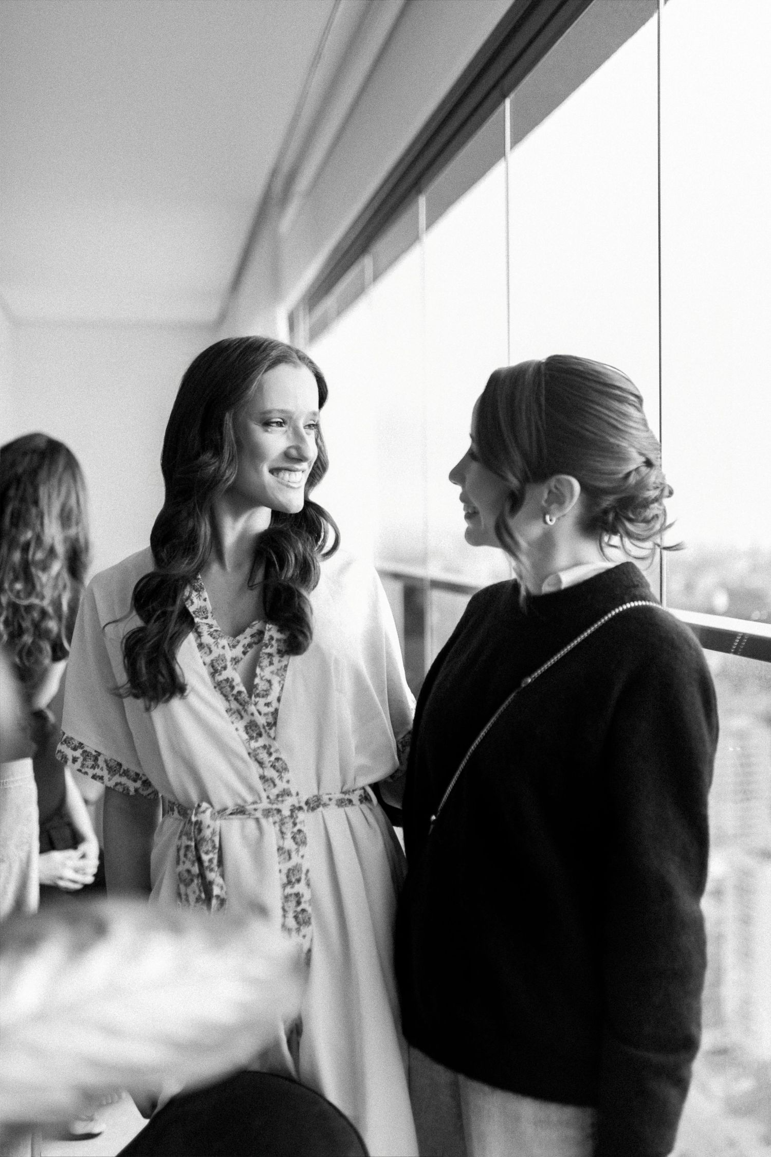 Black and white photo of two women smiling at each other near a window, one wearing a floral robe.
