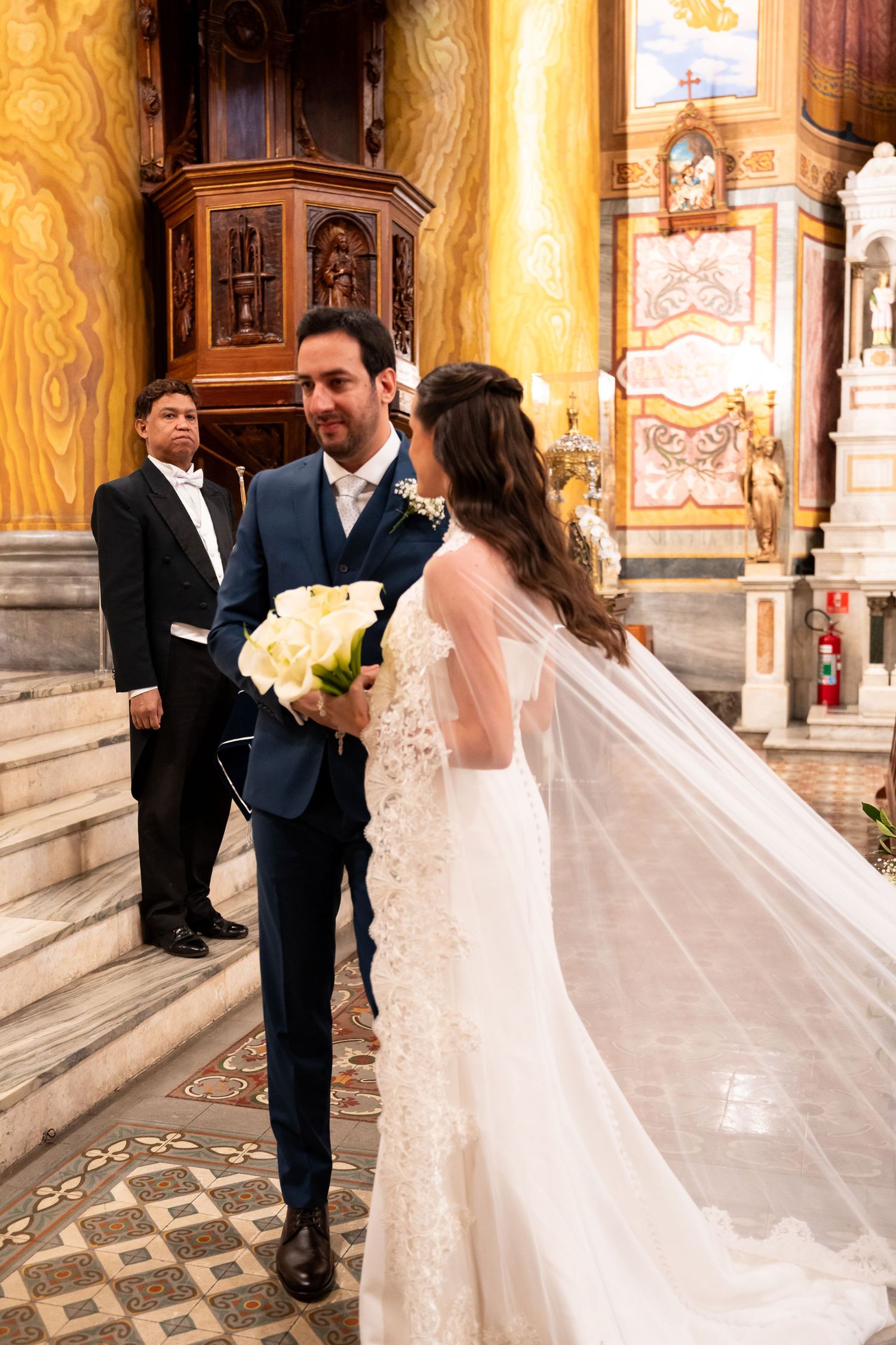 Bride and groom share a romantic moment at church altar, bride holding white calla lily bouquet with veil flowing.