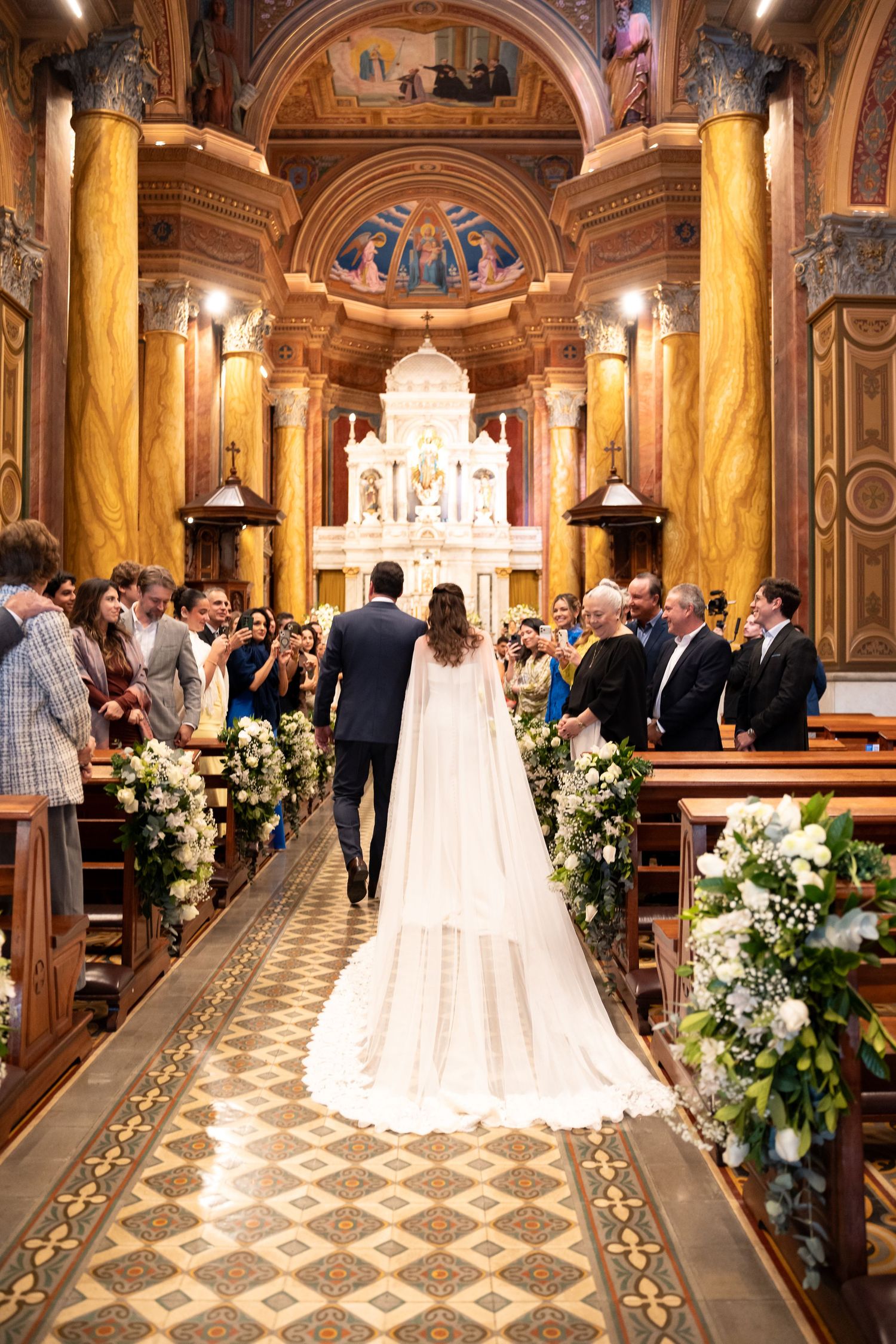 Bride and groom walking down the aisle of an ornate golden church adorned with white floral arrangements.
