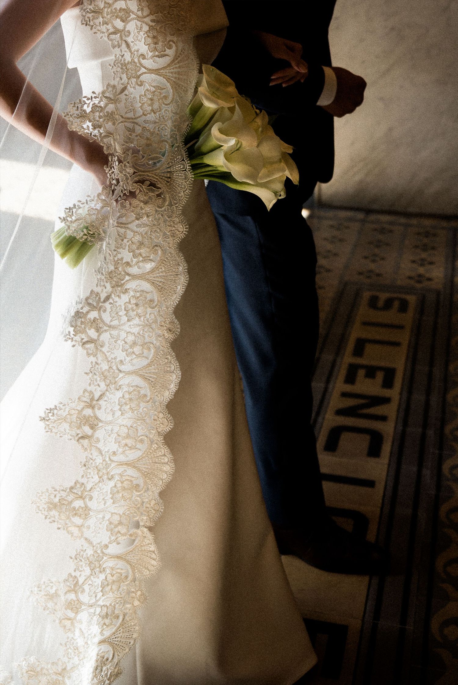 Bride in elegant lace wedding gown holding calla lily bouquet stands beside groom near a Silencio sign.