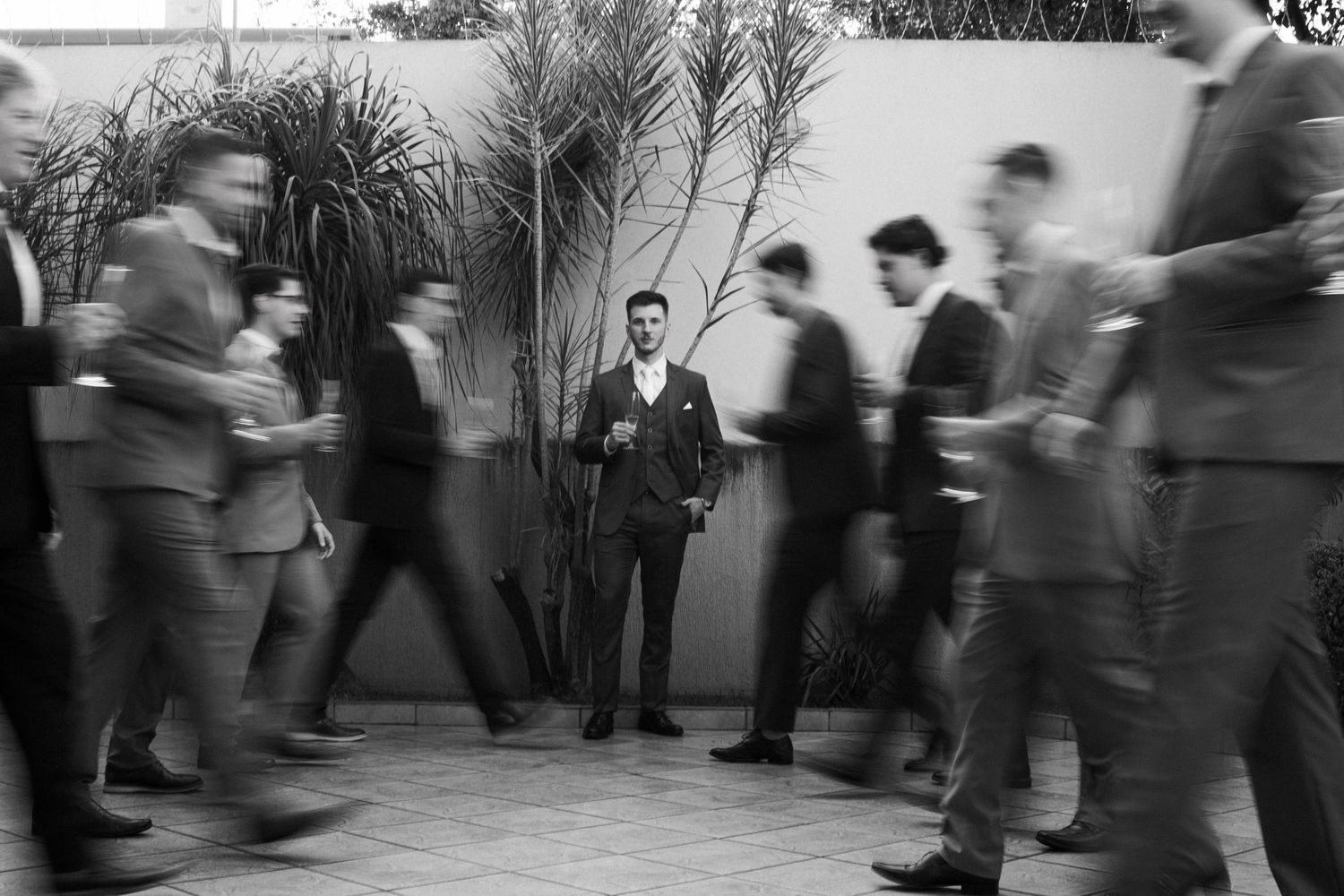 Black and white photo of groom standing still while blurred wedding guests move around him outdoors.