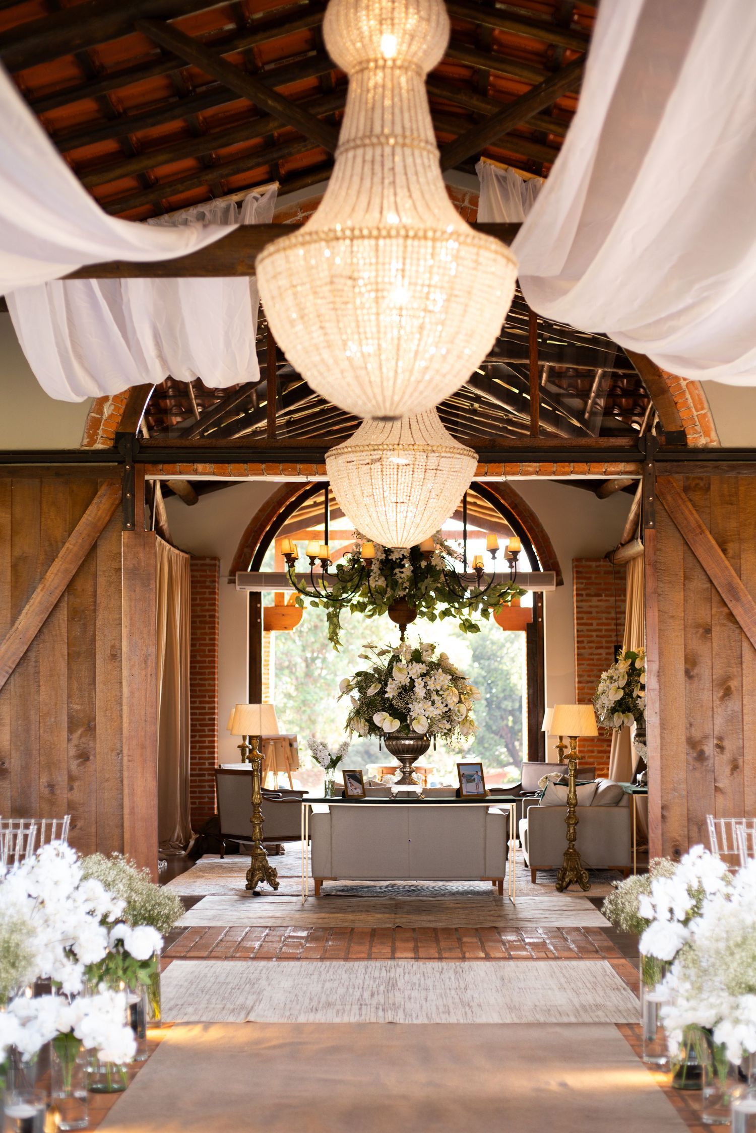 Elegant barn wedding venue with crystal chandelier, white draping, floral arrangements, and rustic wooden beams.