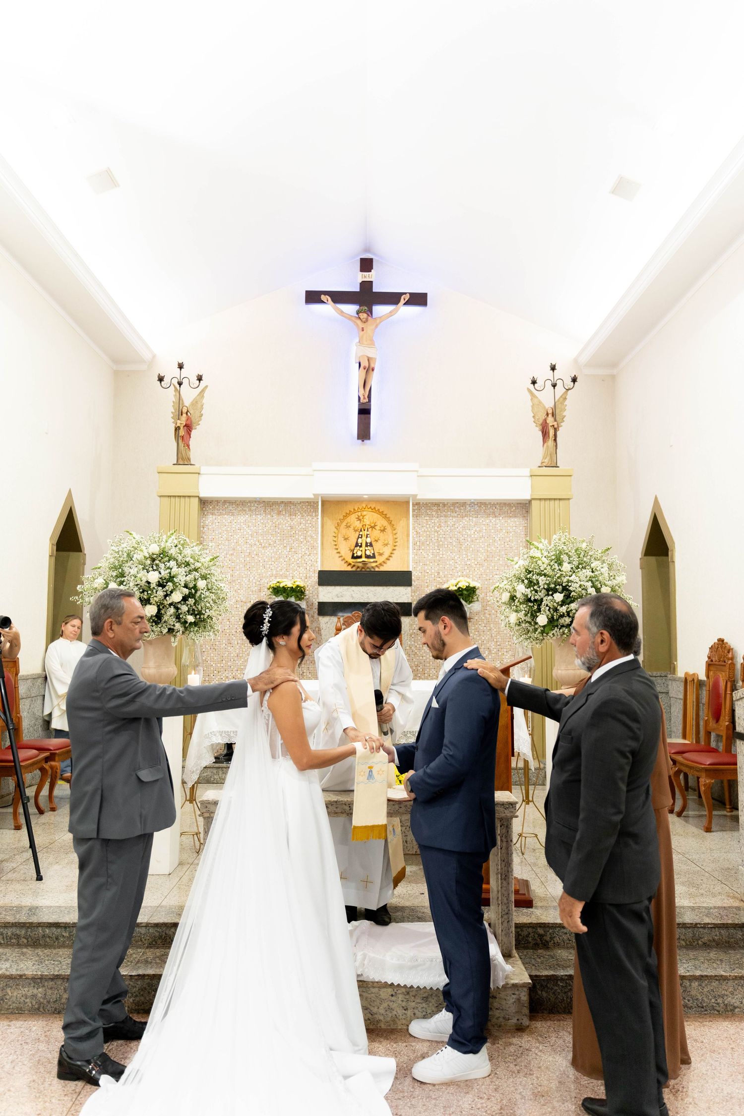 Bride and groom exchange vows at a Catholic church altar, flanked by family, with a crucifix above.