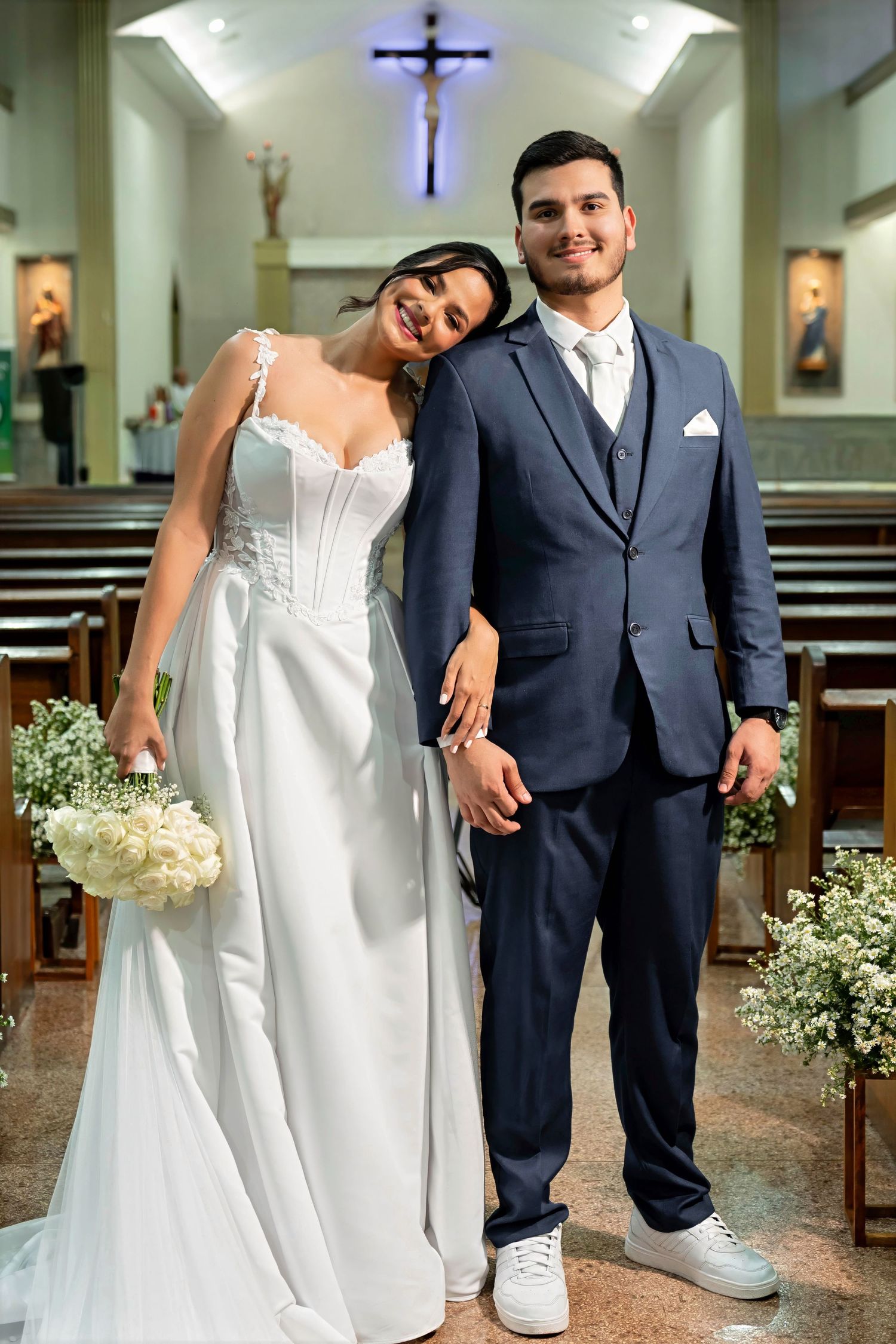 Bride in white gown and groom in navy suit pose together smiling in a church aisle after their wedding ceremony.