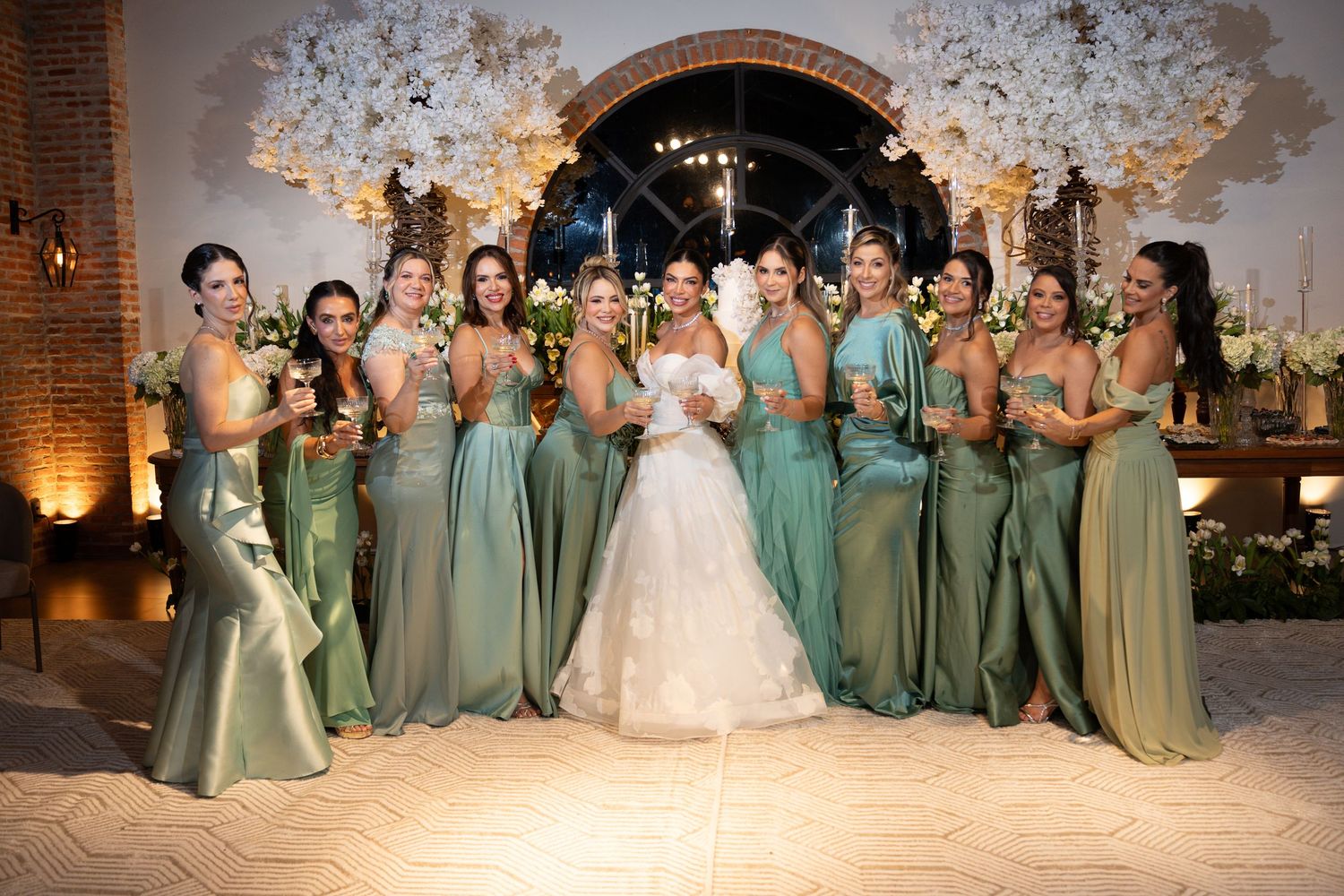 Bride in white ball gown poses with ten bridesmaids in sage green dresses at elegant floral wedding venue.