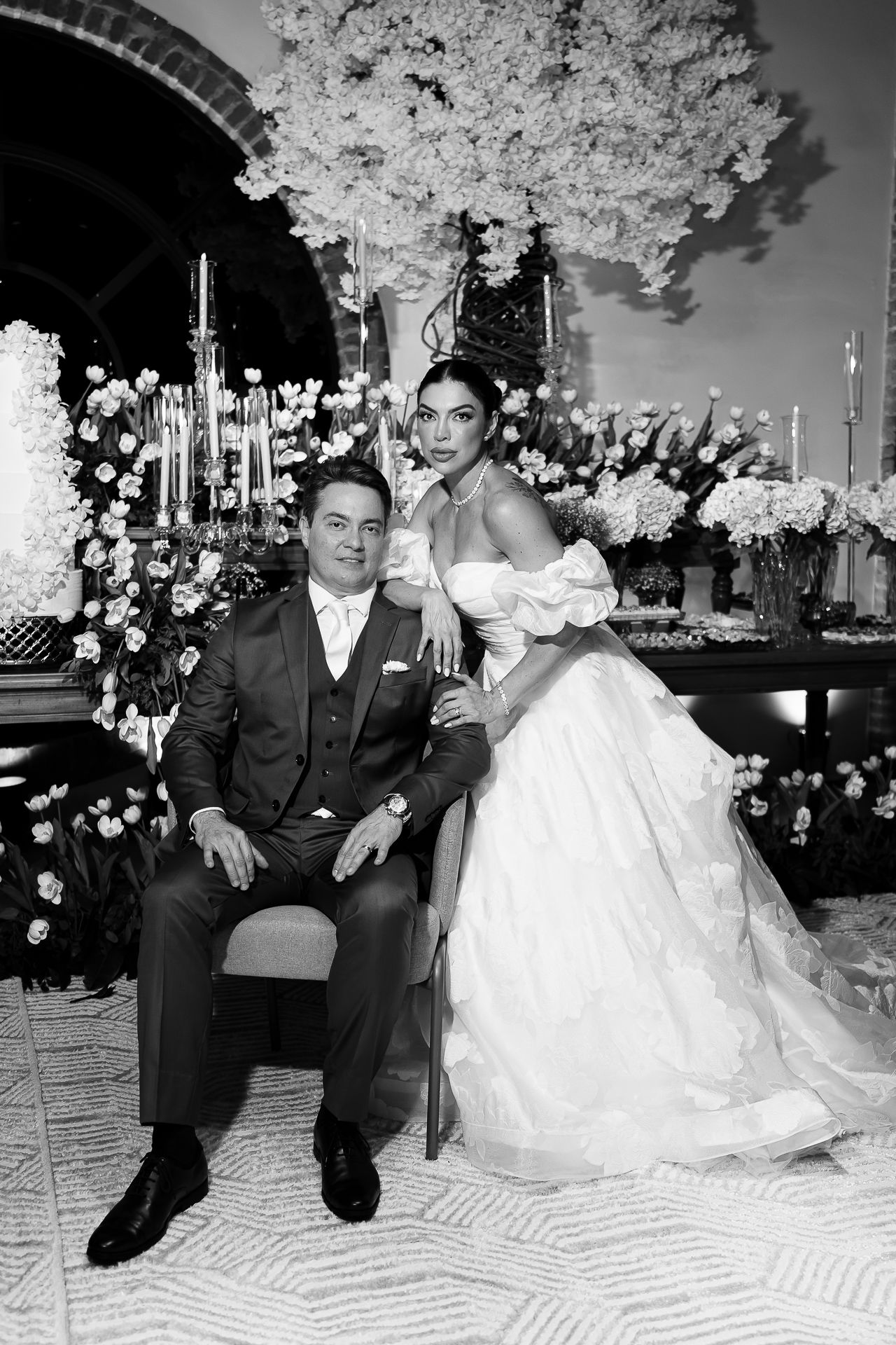 Black and white wedding photo of bride in off-shoulder gown and groom in suit posing before floral backdrop.