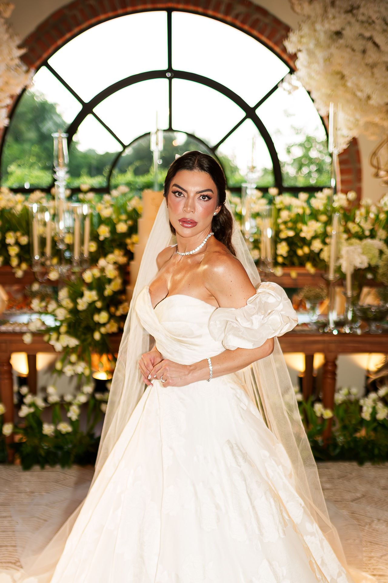 Elegant bride in strapless white ball gown with veil and pearl necklace poses before floral arch backdrop.