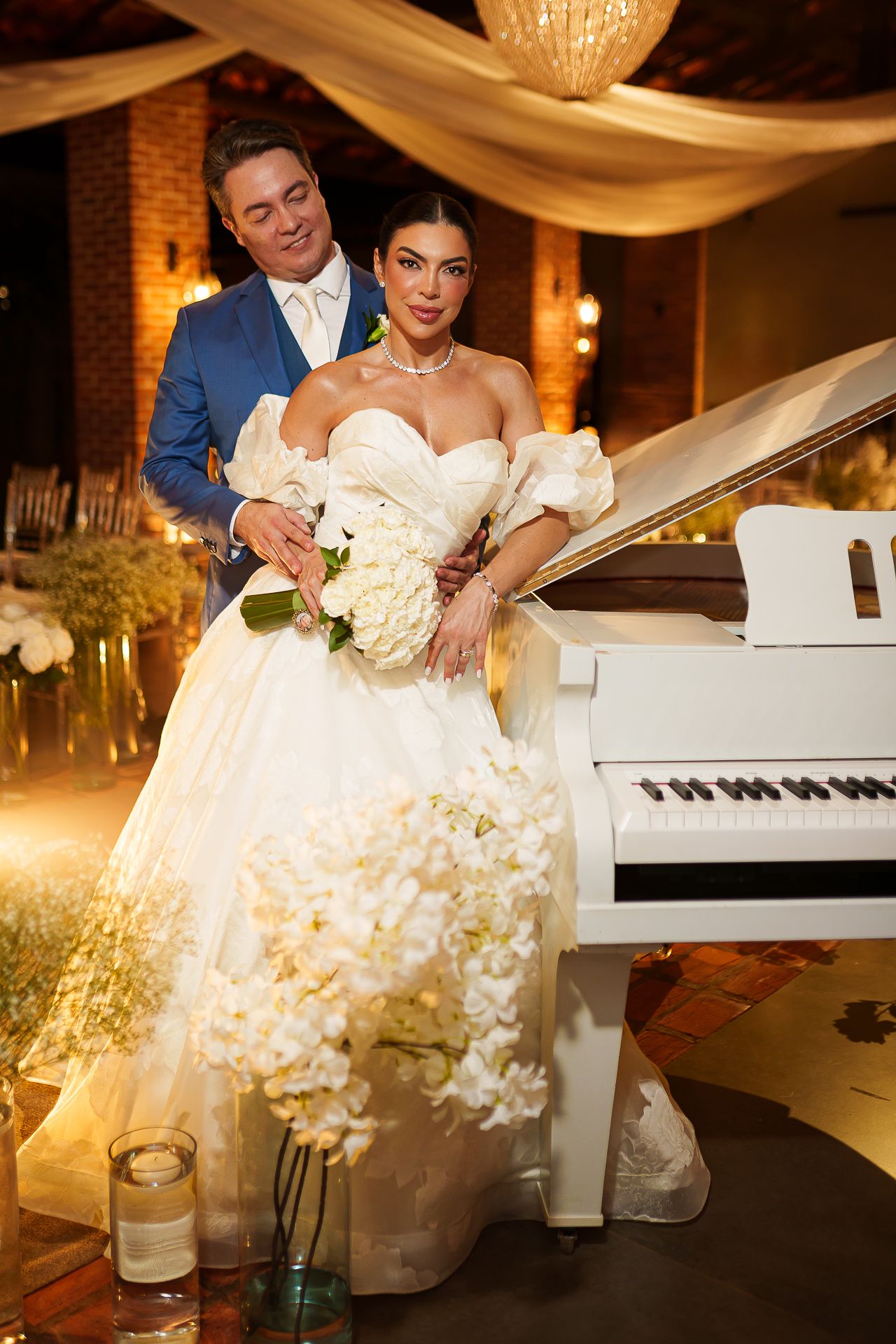 Elegant bride in off-shoulder ball gown holding white bouquet poses with groom beside a white grand piano at wedding.