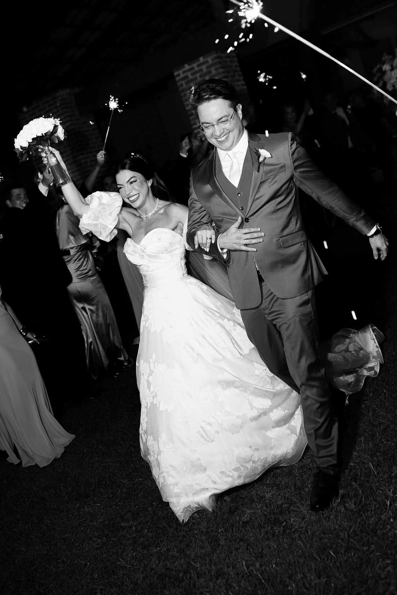 Black and white photo of a joyful newlywed couple running through sparklers at their wedding reception at night.