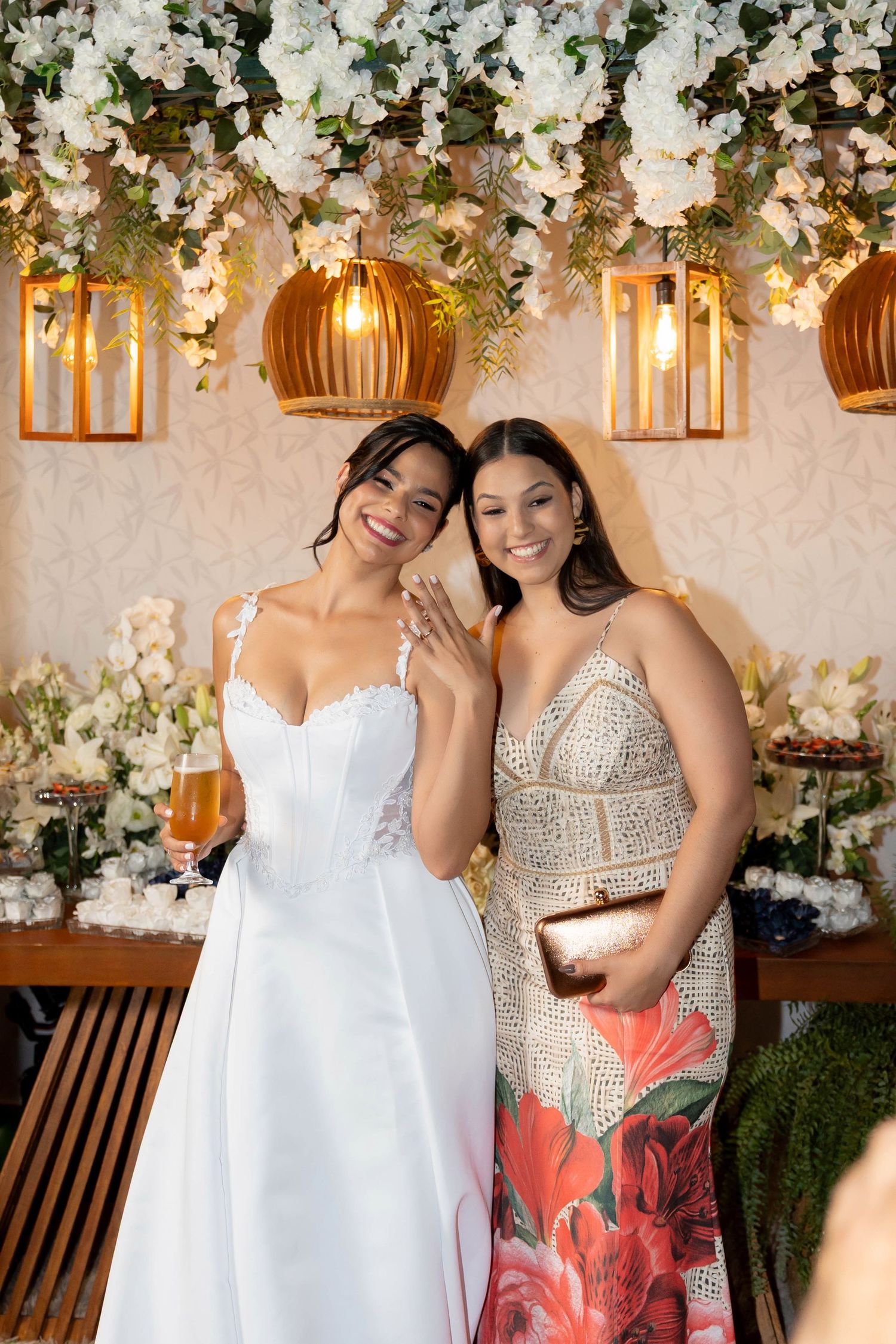 Bride in white gown shows off ring with guest in floral dress at elegant wedding reception with flower decor.
