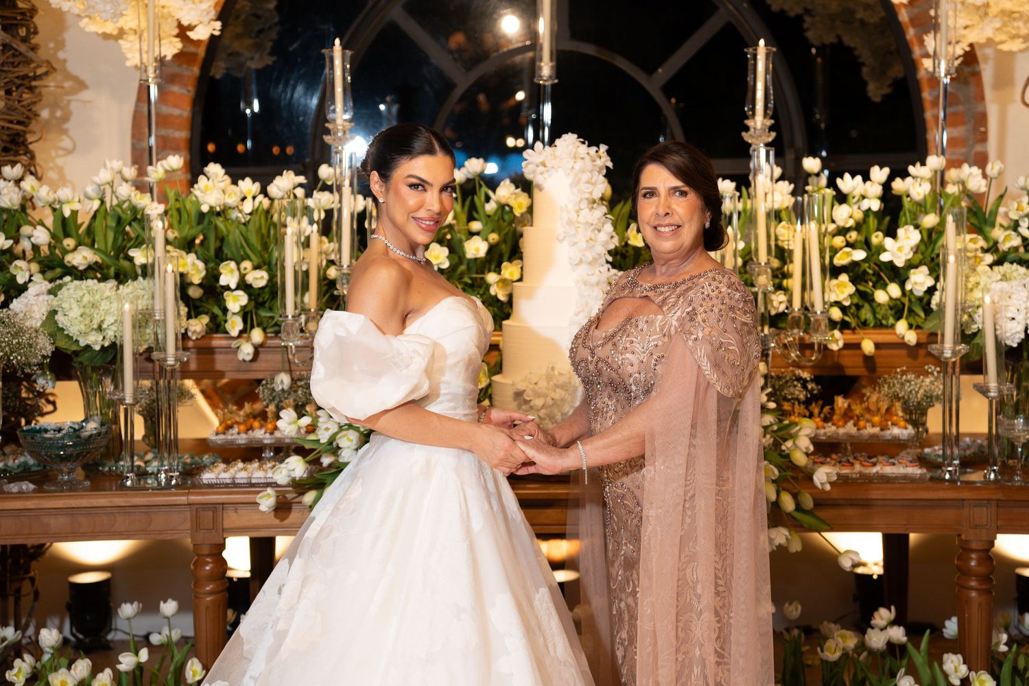 Bride in white gown and older woman in beige dress cutting elegant white wedding cake at floral-decorated reception.