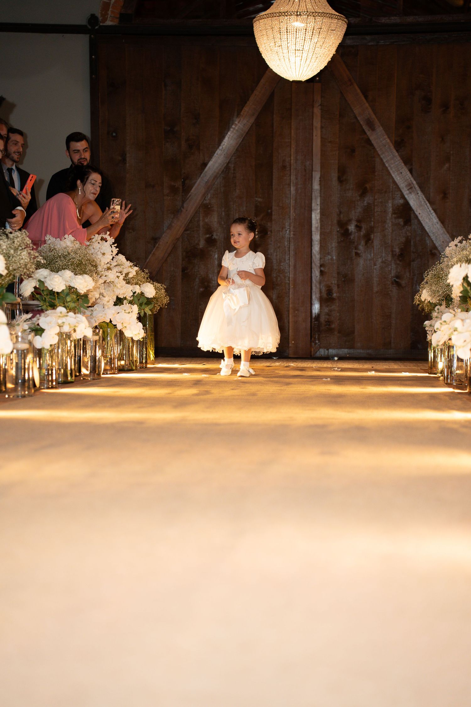 A flower girl in a white dress walks down a floral-lined wedding aisle in a rustic barn venue.