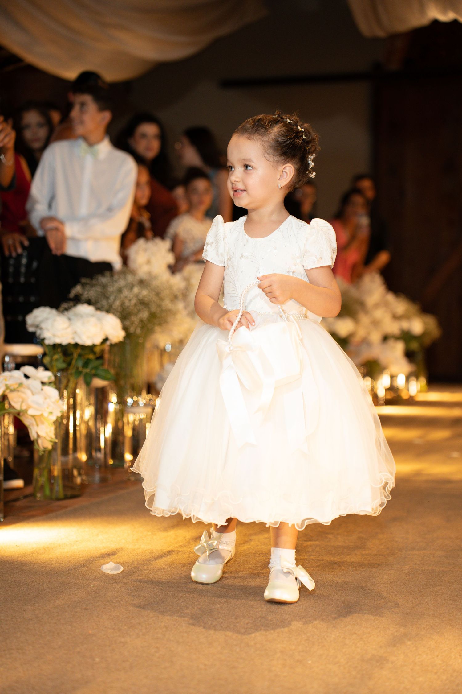 Adorable flower girl in white tulle dress walking down wedding aisle, smiling, with floral arrangements and guests in background.