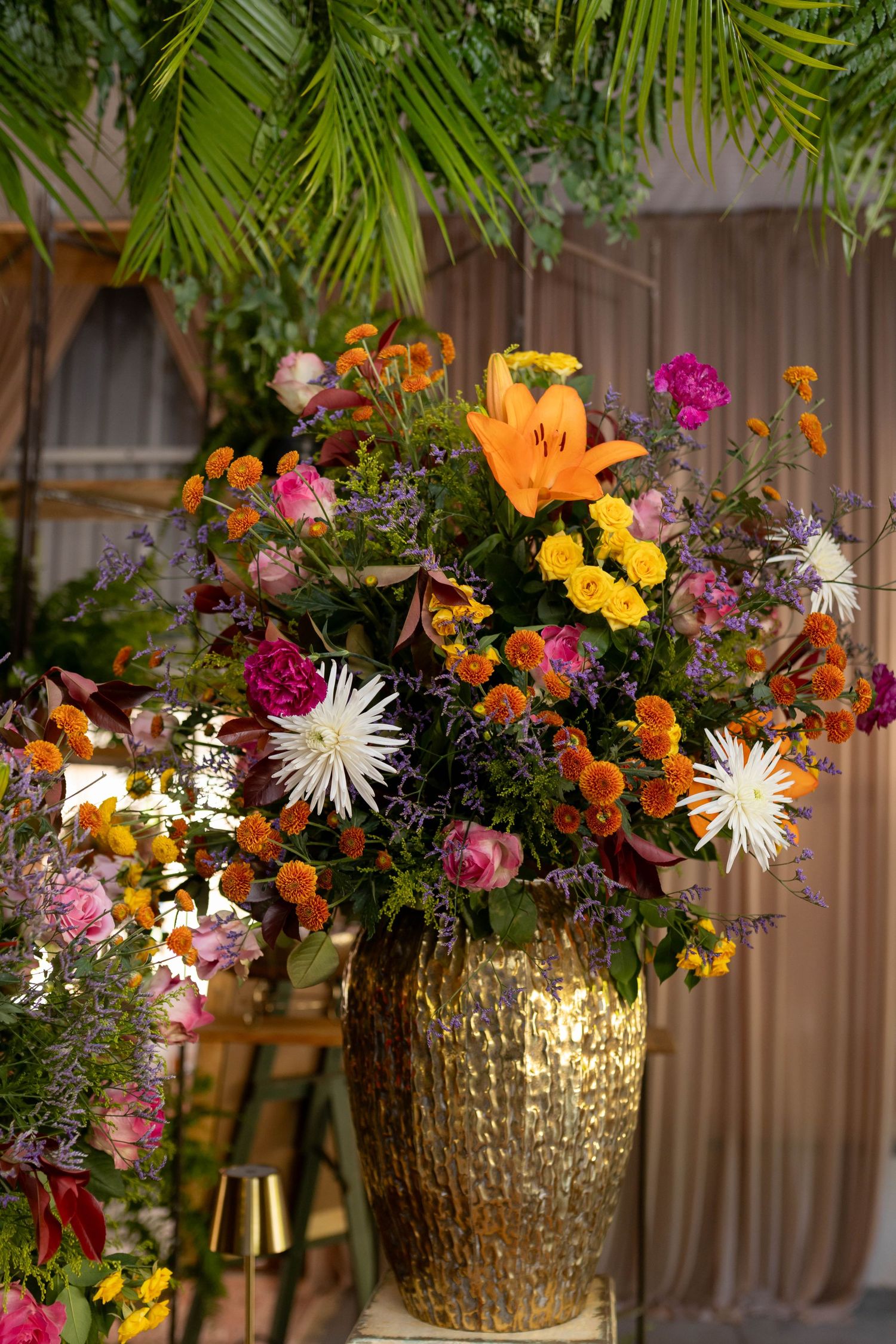 Lush floral arrangement in gold vase with orange lilies, pink roses, and colorful blooms against draped backdrop.