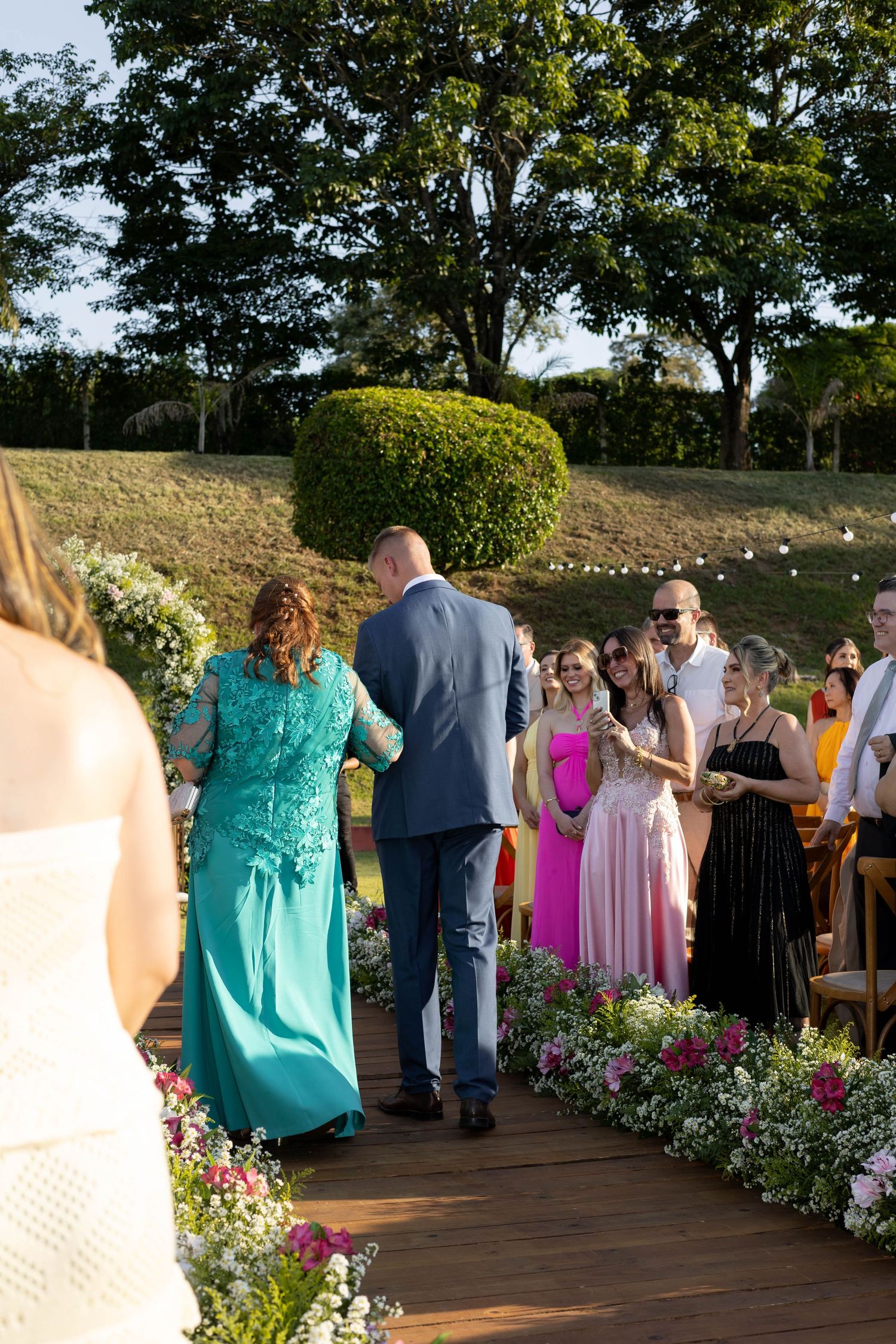 Outdoor wedding ceremony on wooden deck with guests in colorful formal attire lining the floral aisle.