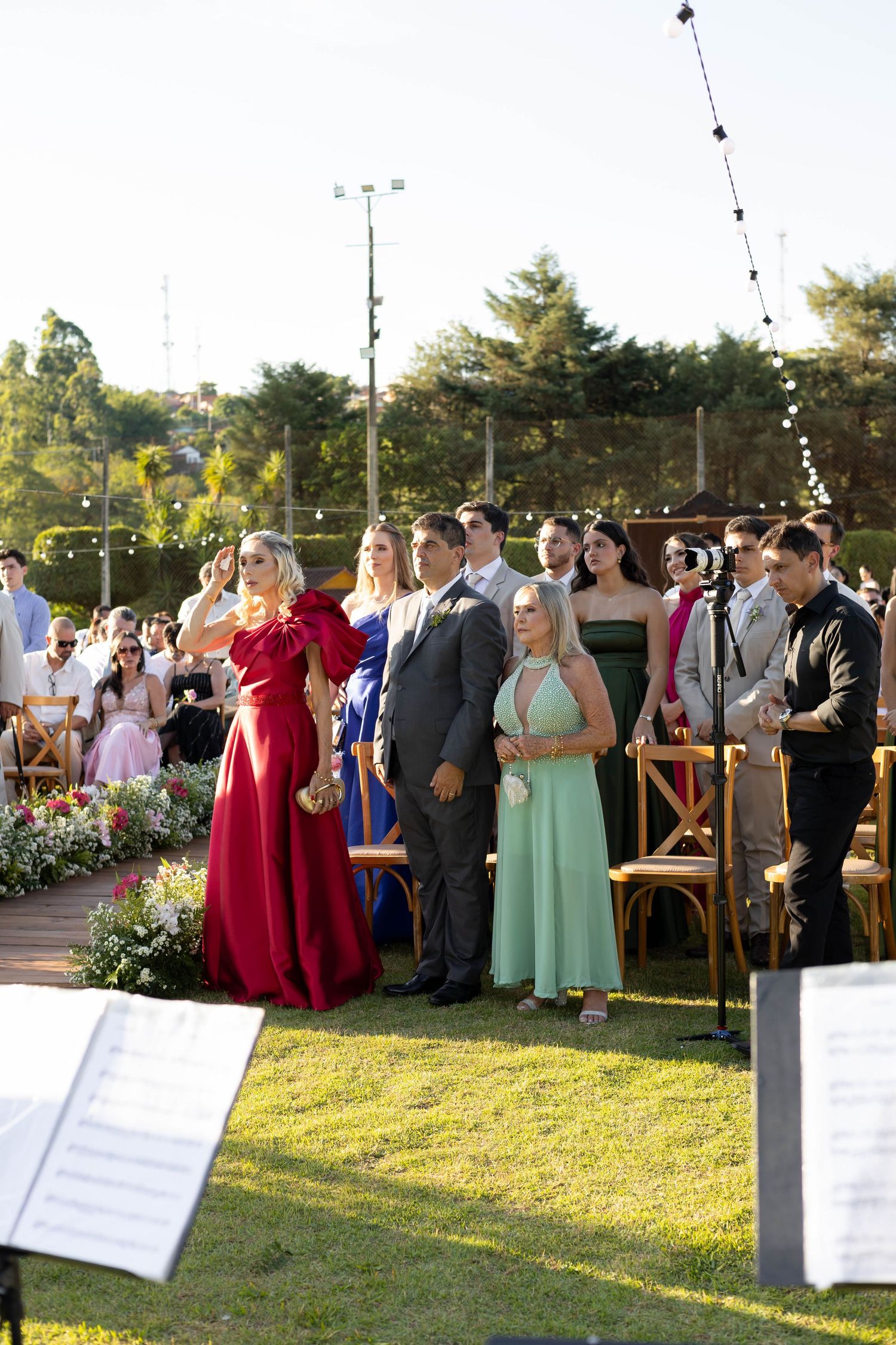 Guests in formal attire stand outdoors at an elegant garden wedding ceremony on a sunny day.