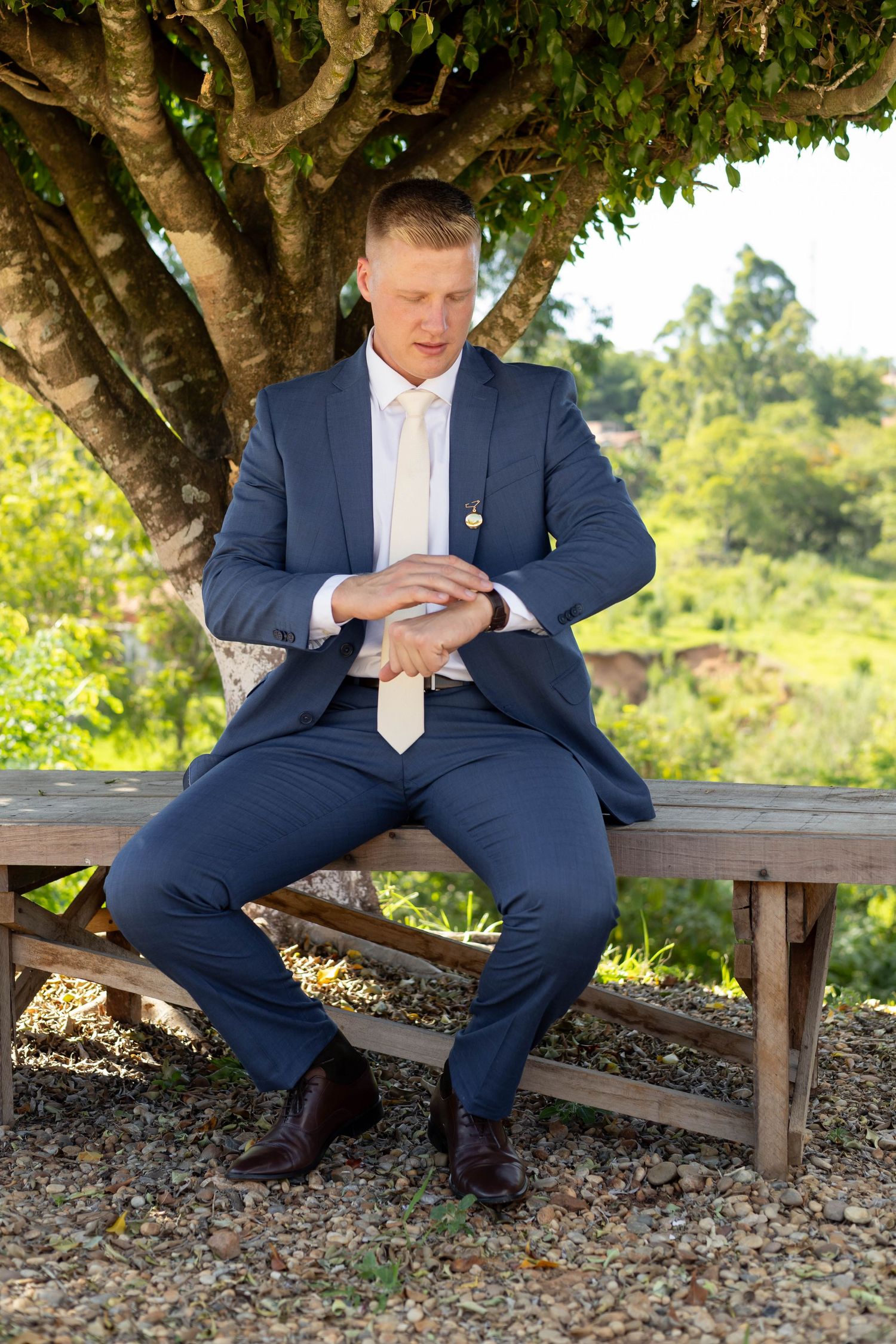 A man in a navy blue suit and tie sits on a wooden bench outdoors, looking down at his hands.
