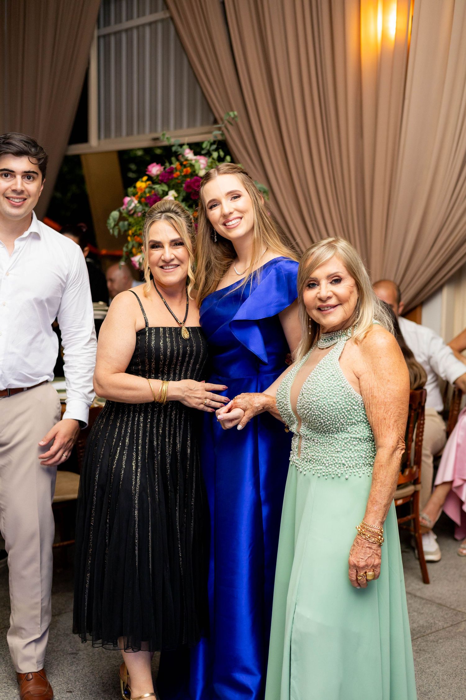 Three women in elegant gowns pose together at a formal event, with a young man in white shirt nearby.