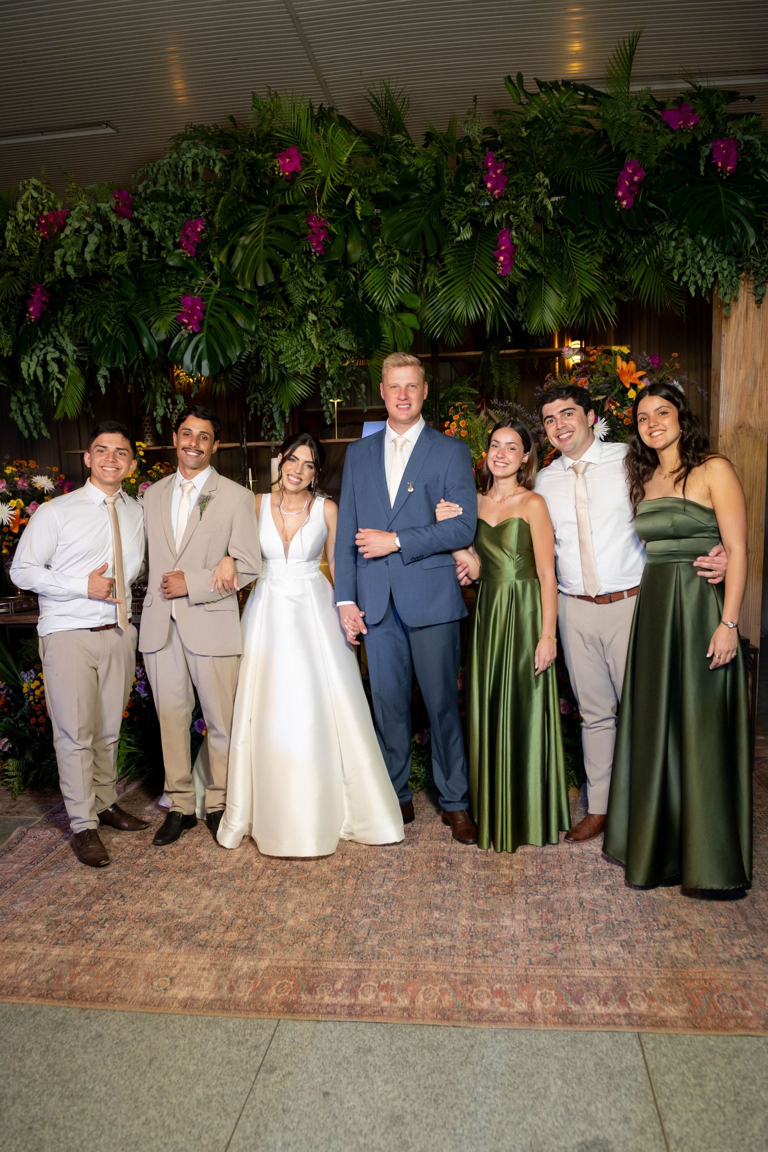 Wedding party posing in front of lush tropical greenery, bride in white gown with groom and guests in formal attire.