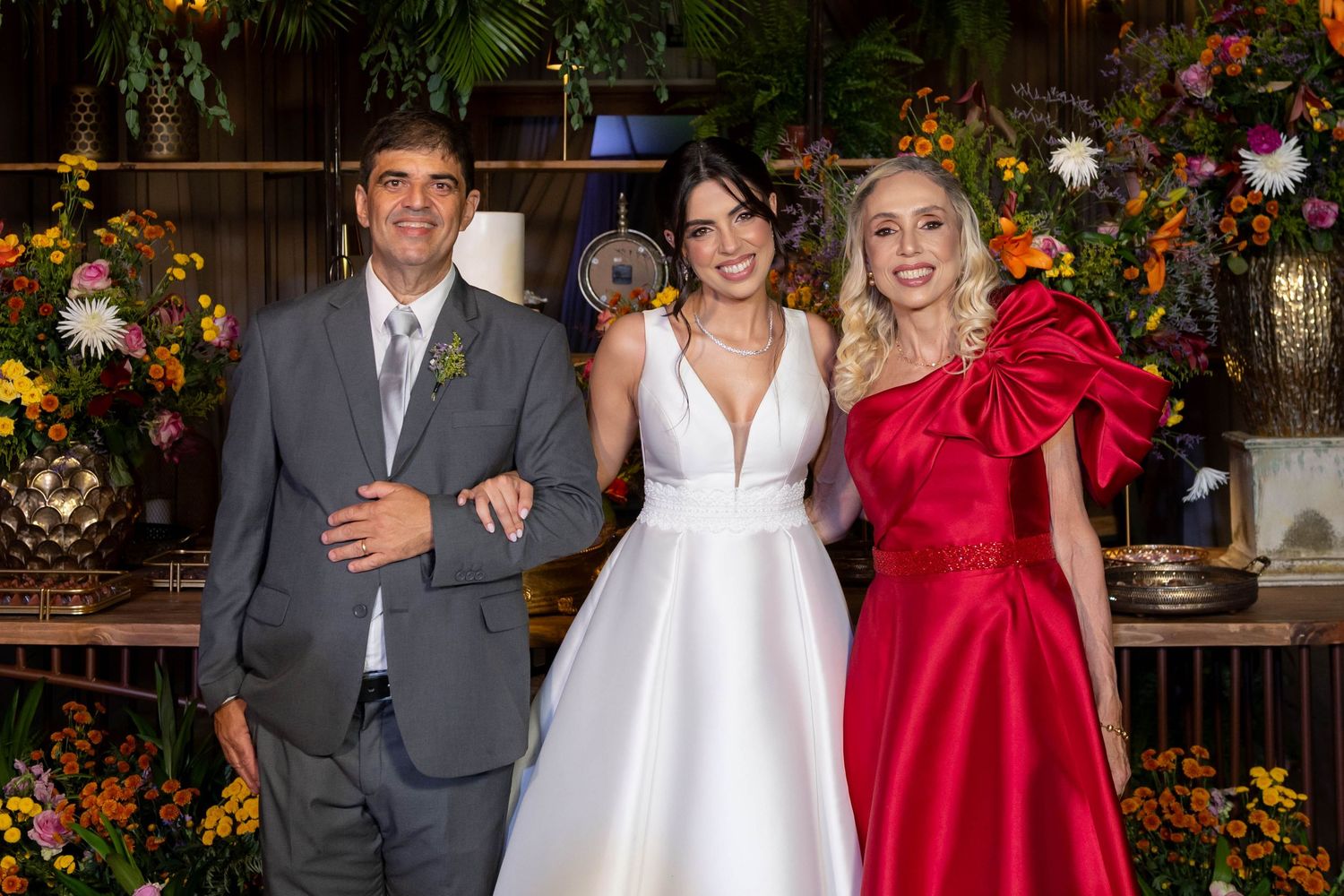 Bride in white gown poses with man in gray suit and woman in red dress at floral wedding reception.