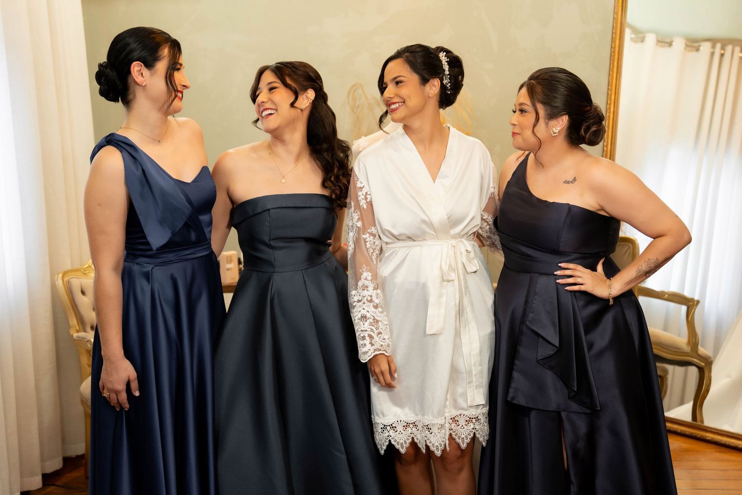 Bride in white robe laughing with three bridesmaids in navy blue dresses during wedding preparations.