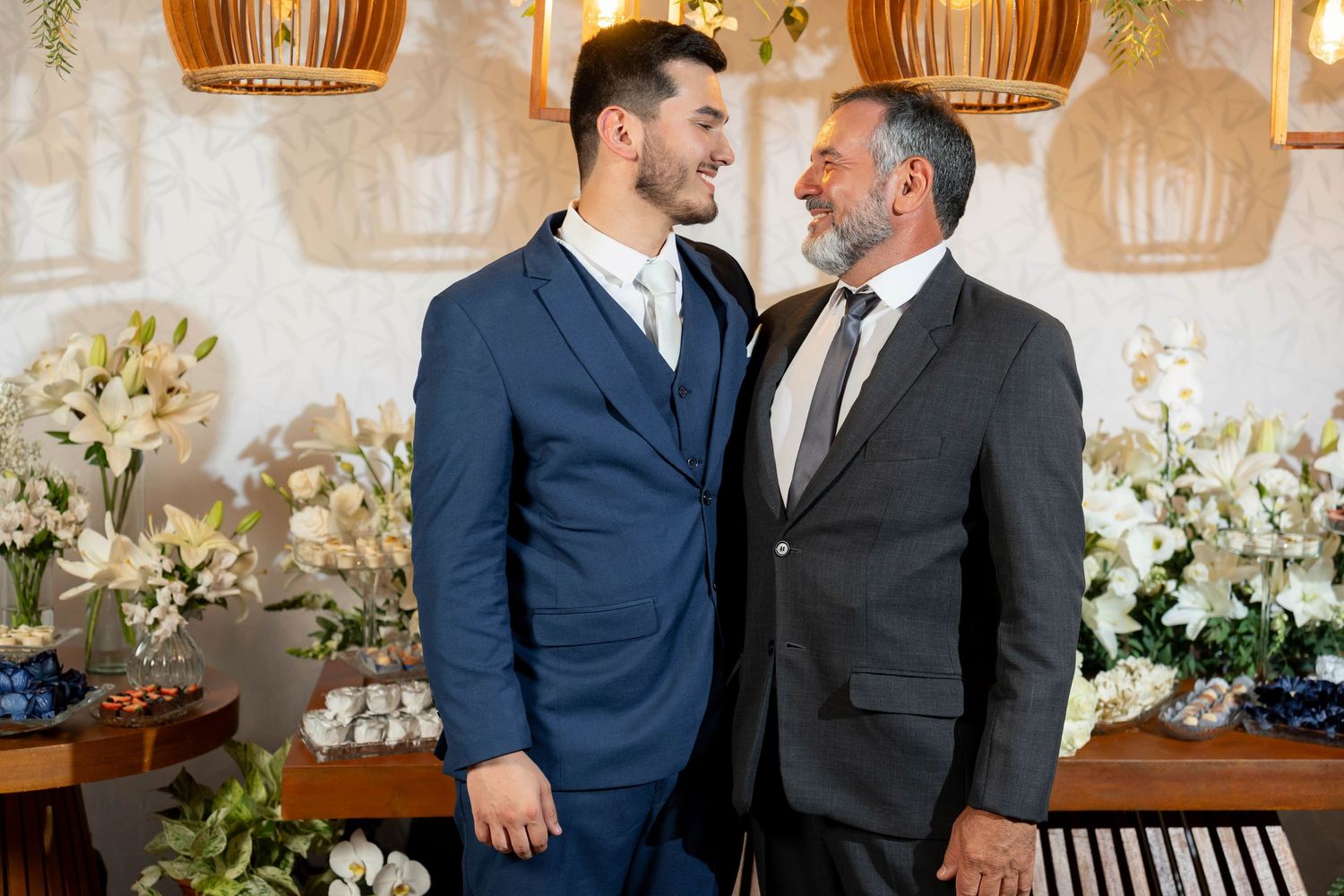 Two men in suits sharing a tender moment at a floral wedding ceremony venue.