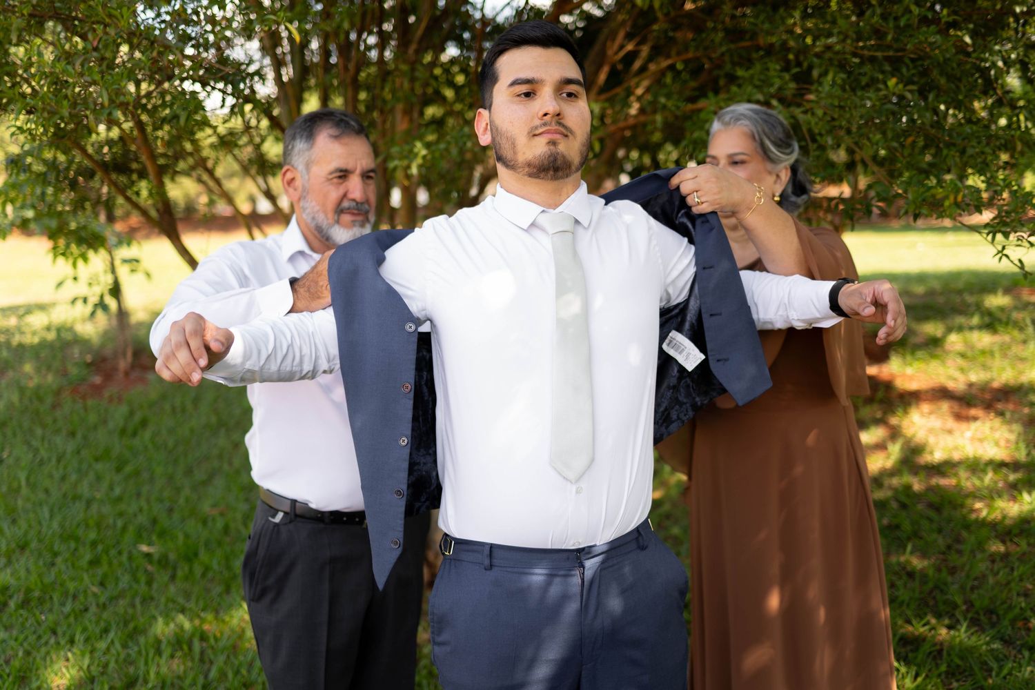 Young man in white shirt and tie gets helped into suit jacket by two older adults outdoors.