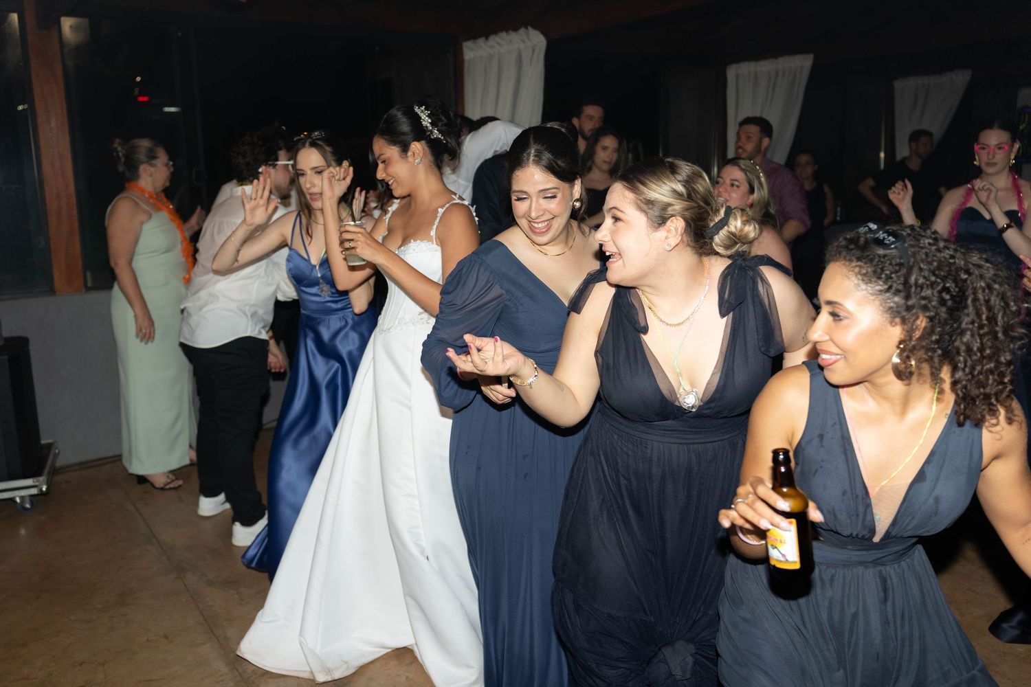 Bride in white gown dancing joyfully with bridesmaids in navy blue dresses at a lively wedding reception.