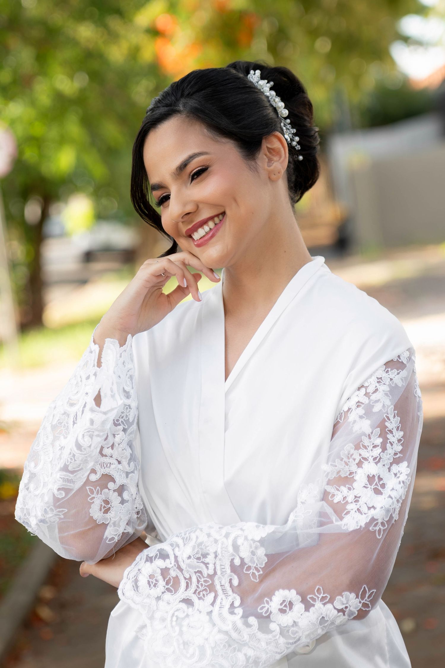 Smiling bride in white lace robe with floral hair accessory posing outdoors among green trees.