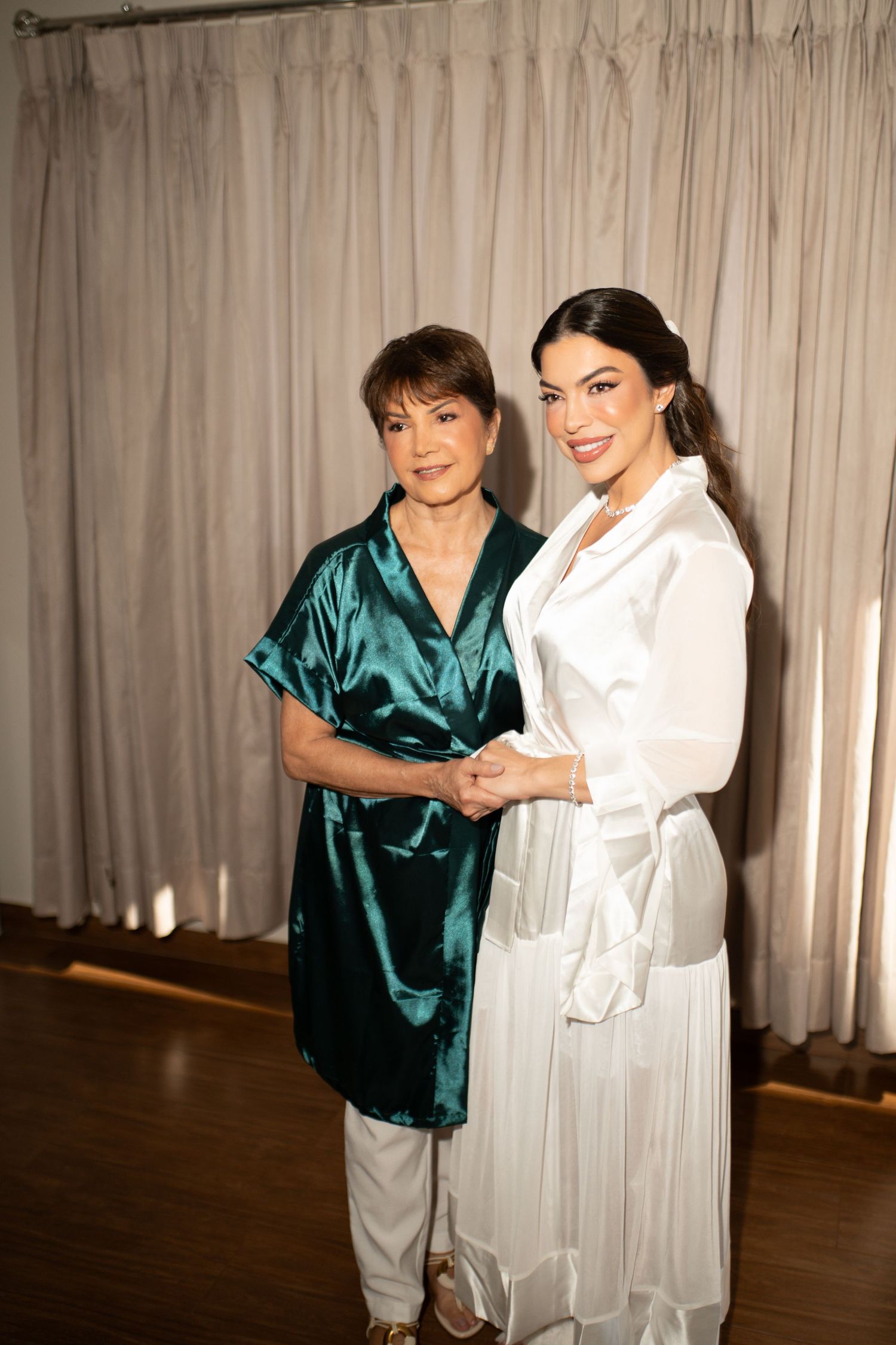 Two women in satin outfits posing together, one in teal and one in white, smiling in an elegant indoor setting.