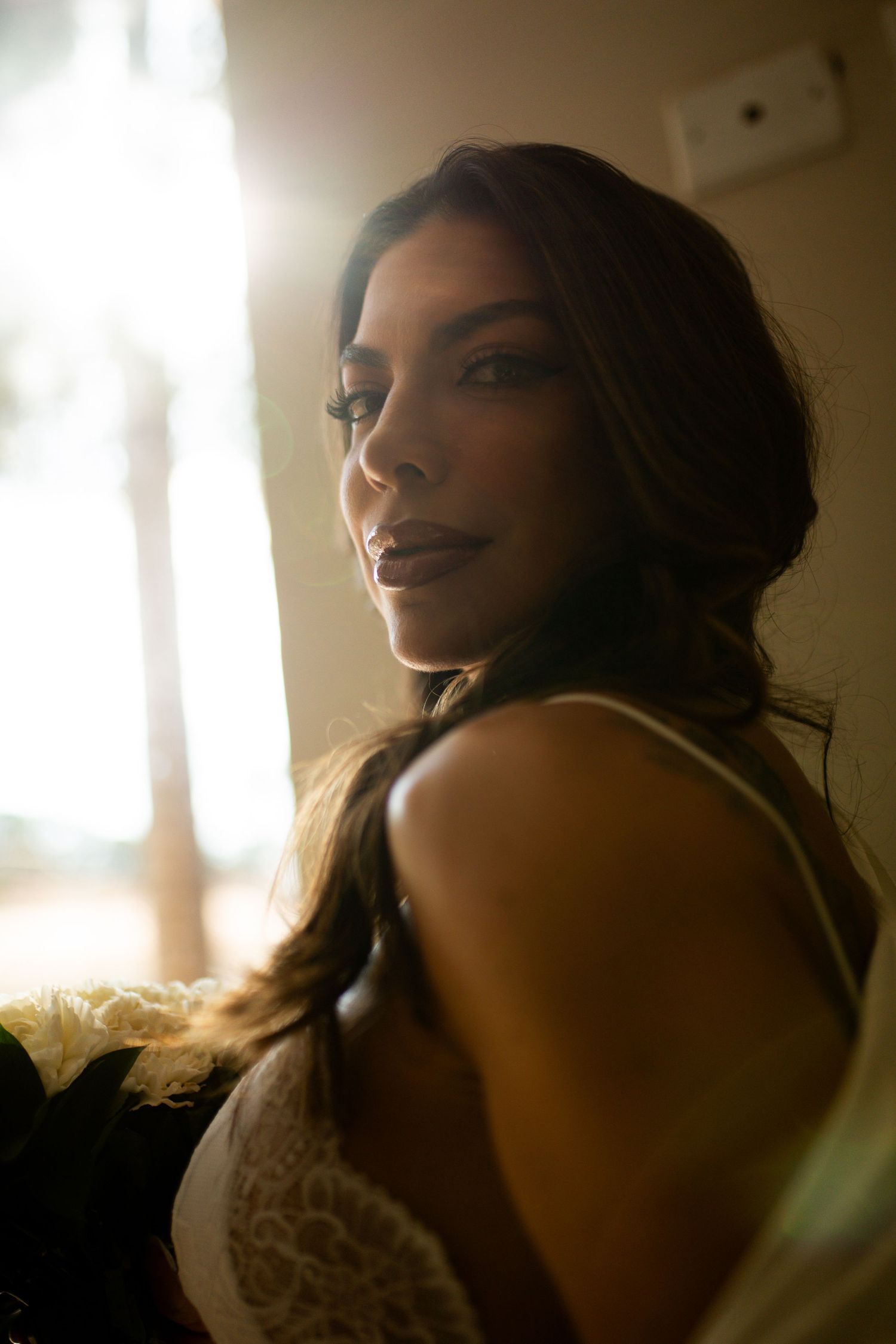 A woman in a lace top gazes over her shoulder in warm backlit indoor light.