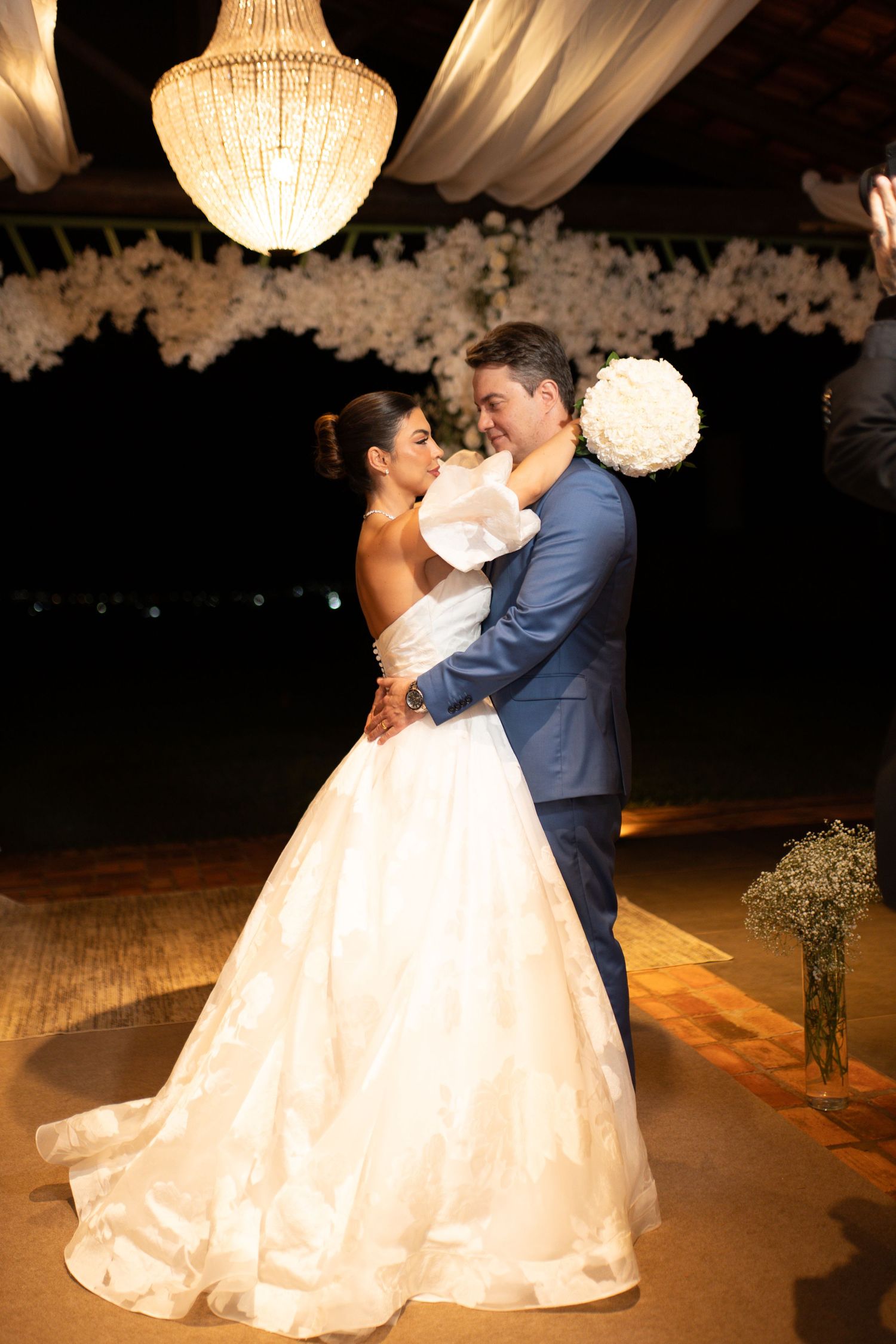 Bride and groom share first dance under crystal chandelier at elegant outdoor wedding reception with white floral backdrop.