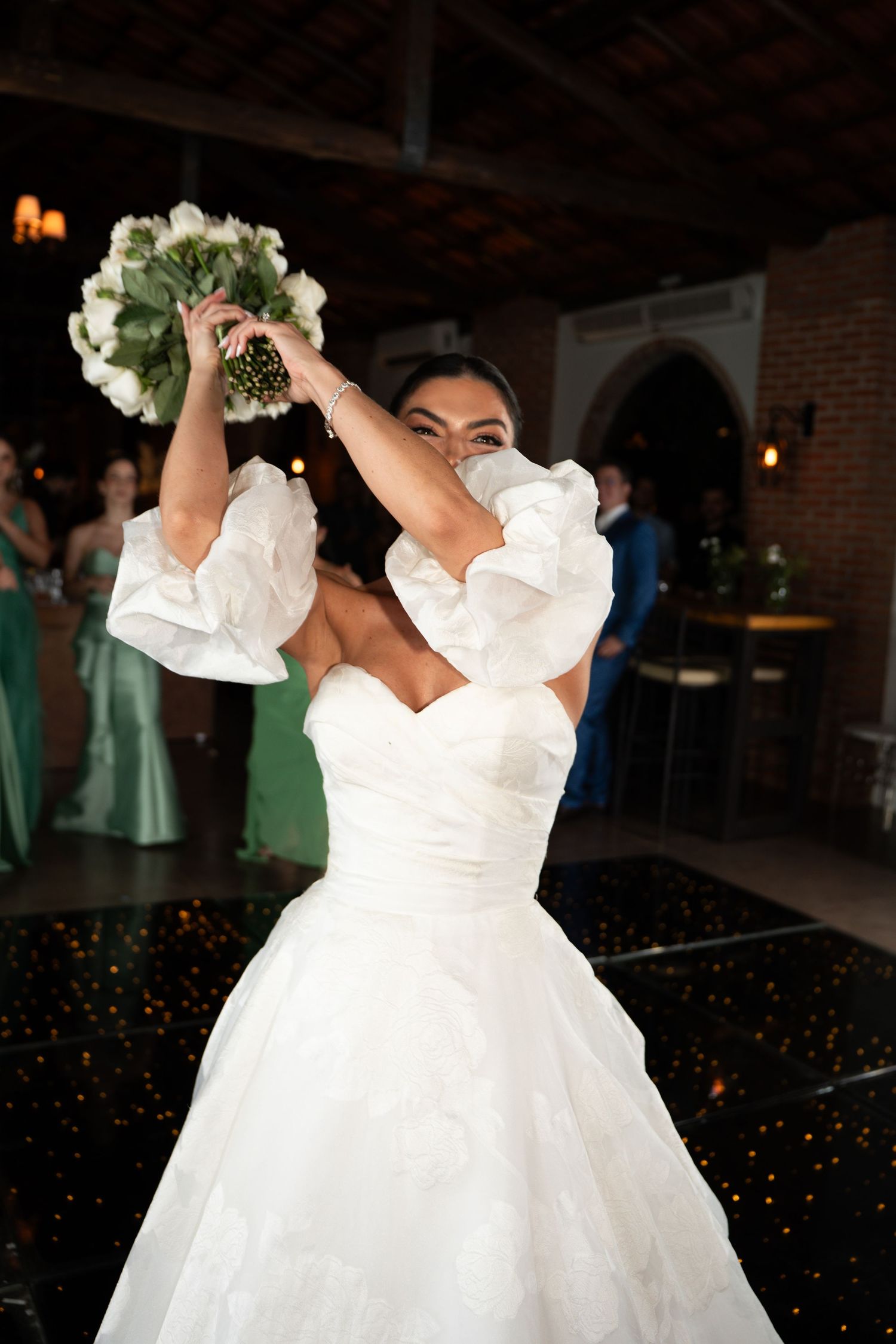 Bride in white ruffled gown tossing bouquet of white flowers at wedding reception.