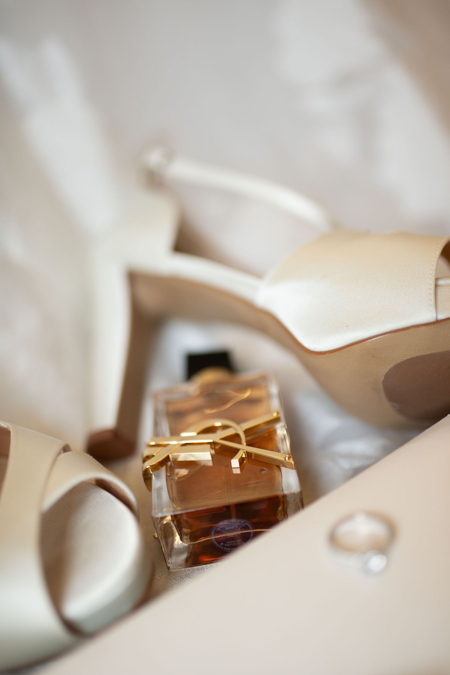 Ivory bridal heels with YSL perfume bottle and diamond ring arranged elegantly on white fabric.